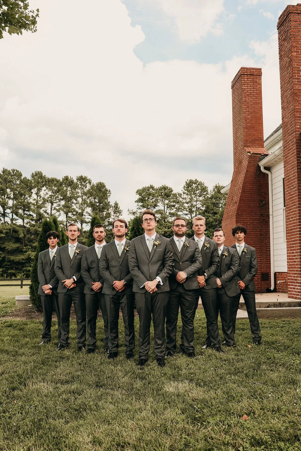 A group of nine men dressed in grey suits with white shirts and light-colored ties, standing outdoors in front of a brick building with large brick chimneys, with trees and a cloudy sky in the background.