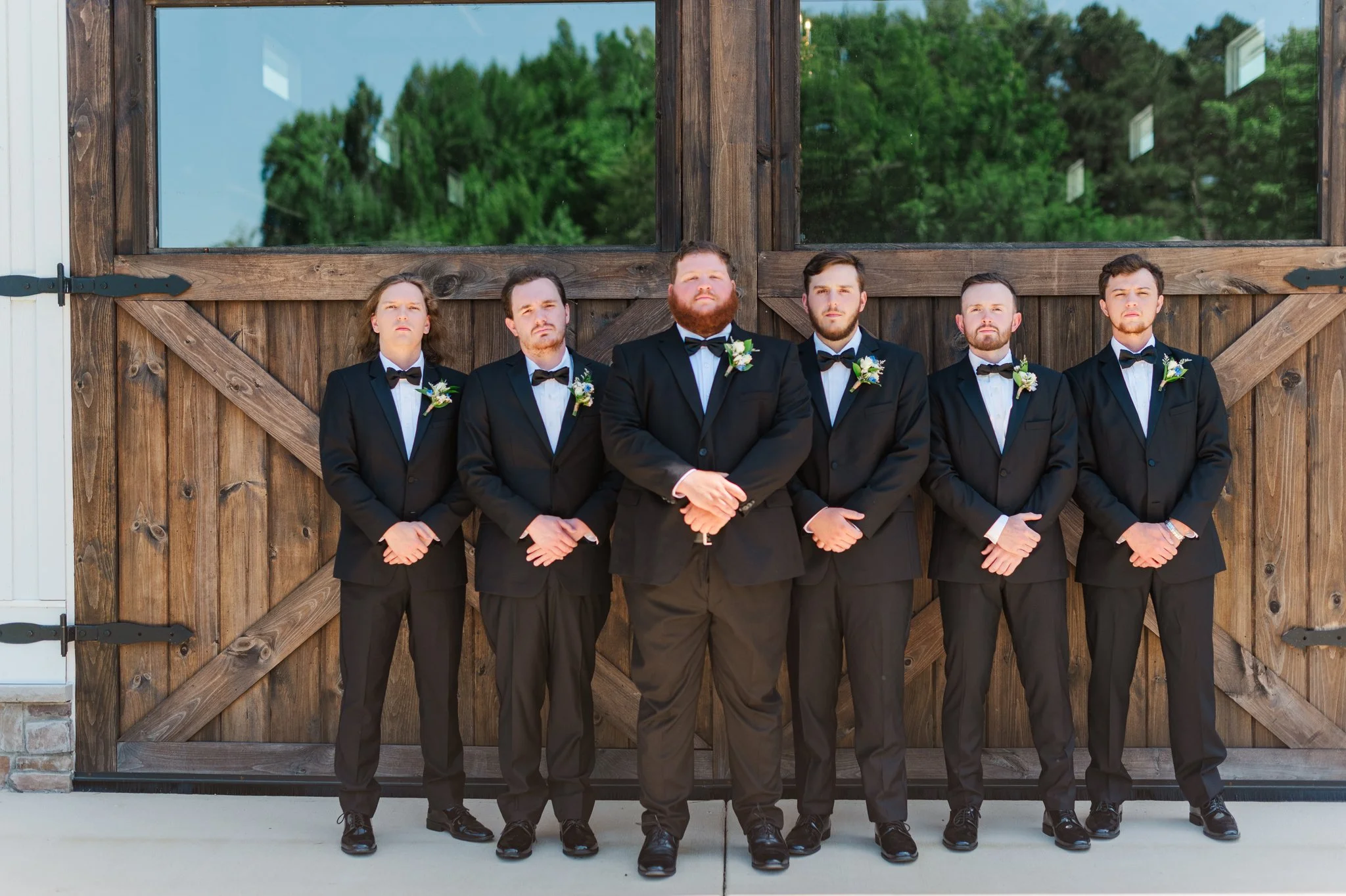 Six men in black tuxedos with bow ties and boutonnières standing in front of a large wooden barn door, with a background of green trees.