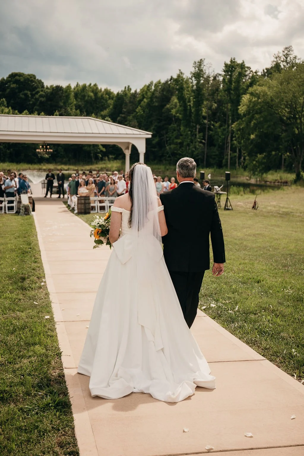 Bride and an older man, likely her father, walking down the aisle at an outdoor wedding ceremony near a lake. The bride is holding a bouquet and wearing a white gown with an off-the-shoulder design and a veil. Guests are gathered in the background un