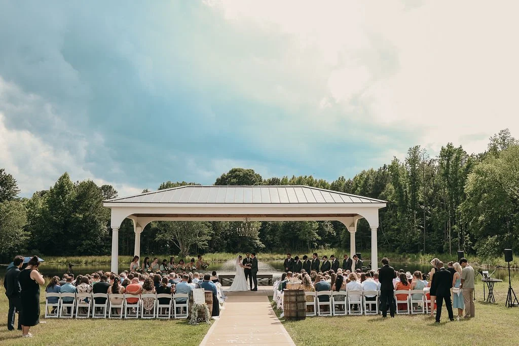 Outdoor wedding ceremony under a gazebo by a lake with guests seated and couple exchanging vows.