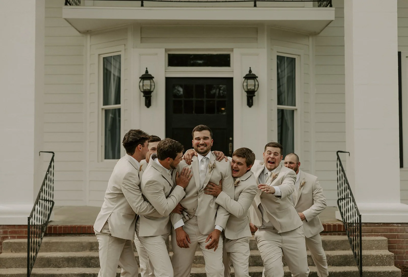 A group of men in beige suits celebrating in front of a white house.