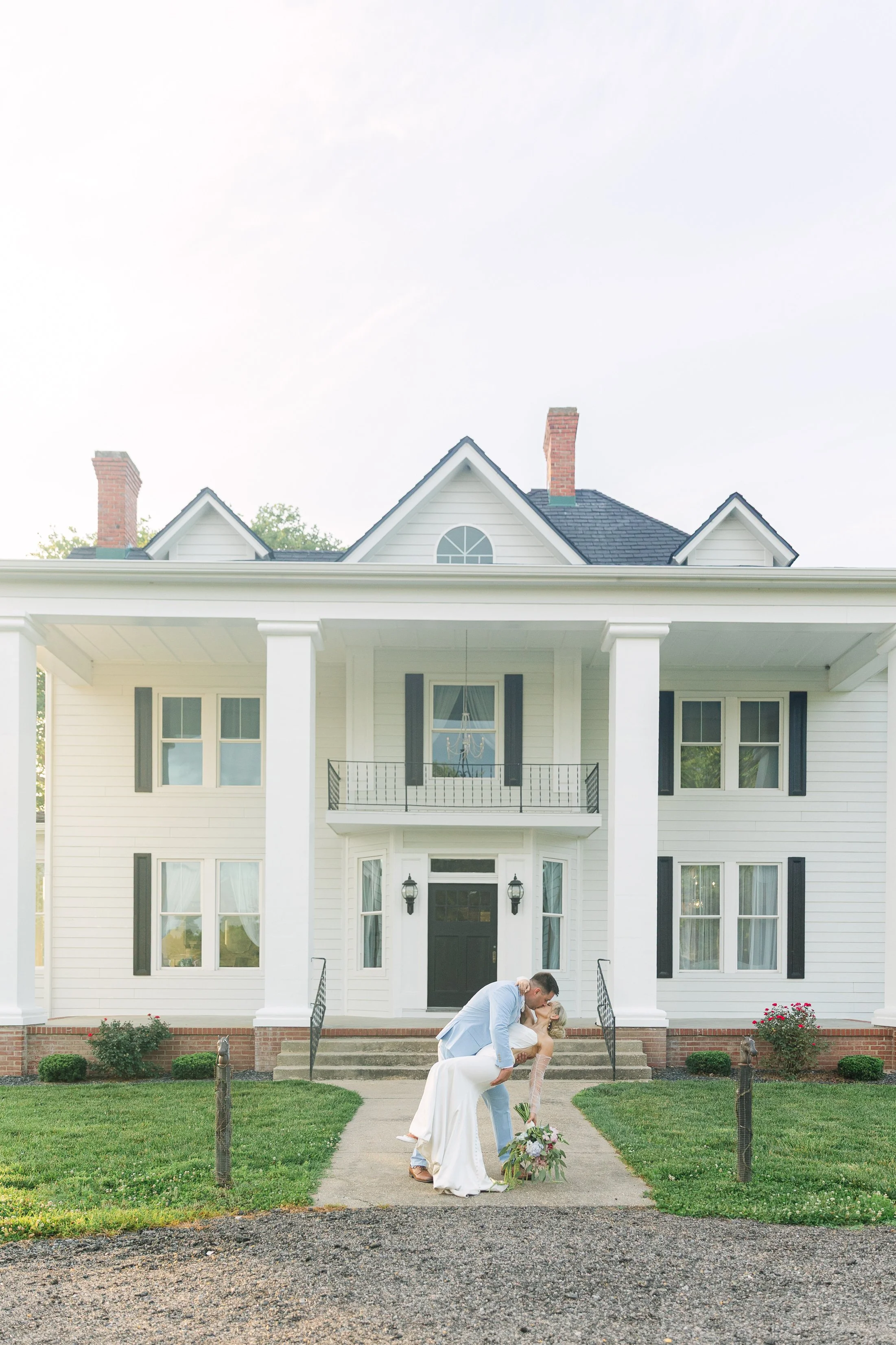 A newlywed couple sharing a kiss in front of a large white house with black shutters. The bride is holding a bouquet of flowers and both are dressed in wedding attire.
