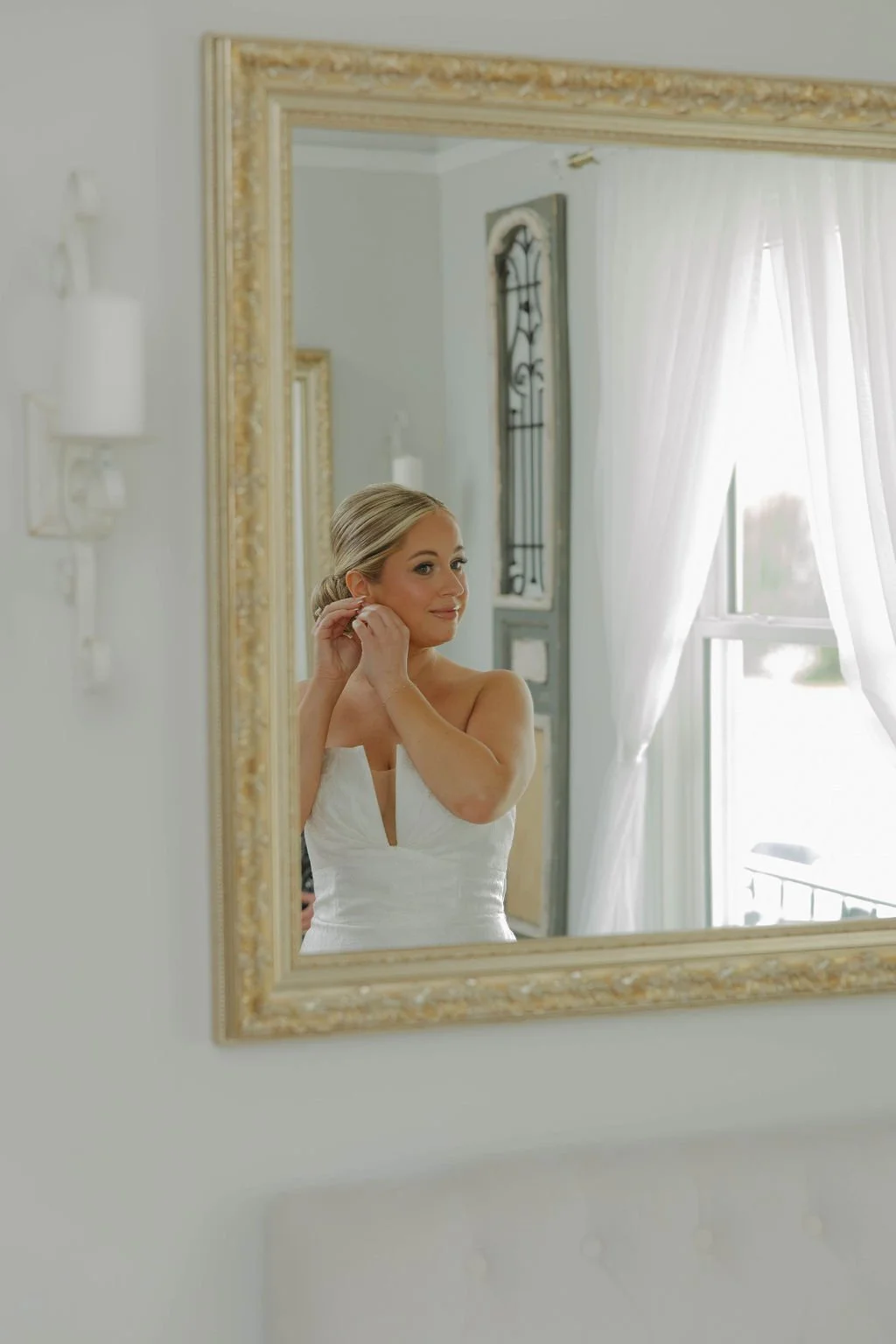A woman in a white dress is seen through a mirror, fixing her earring next to a window with white curtains, in a bright room with ornate decor.