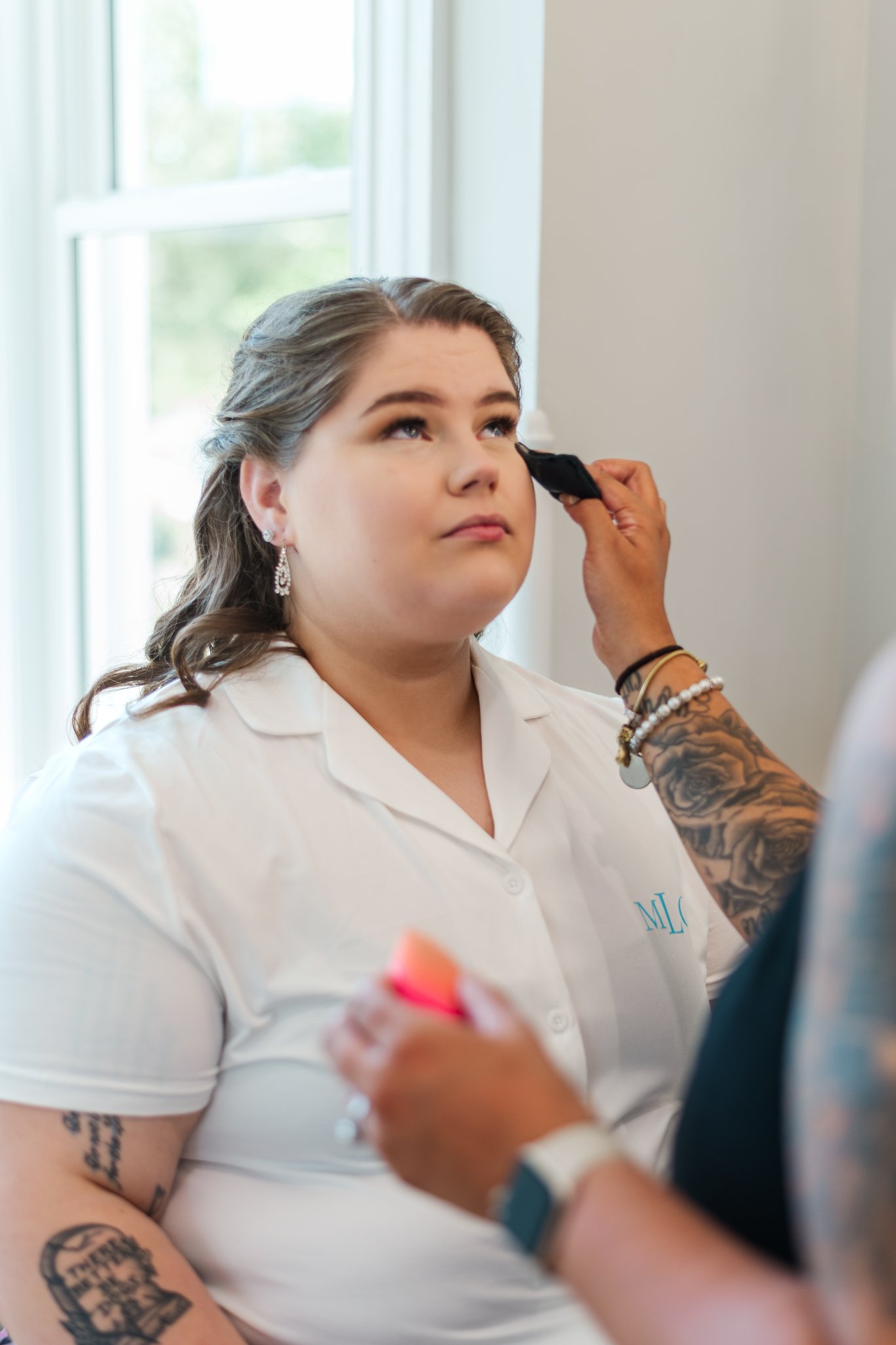 A woman with earrings and tattoos on her arm has makeup applied by a makeup artist near a window.