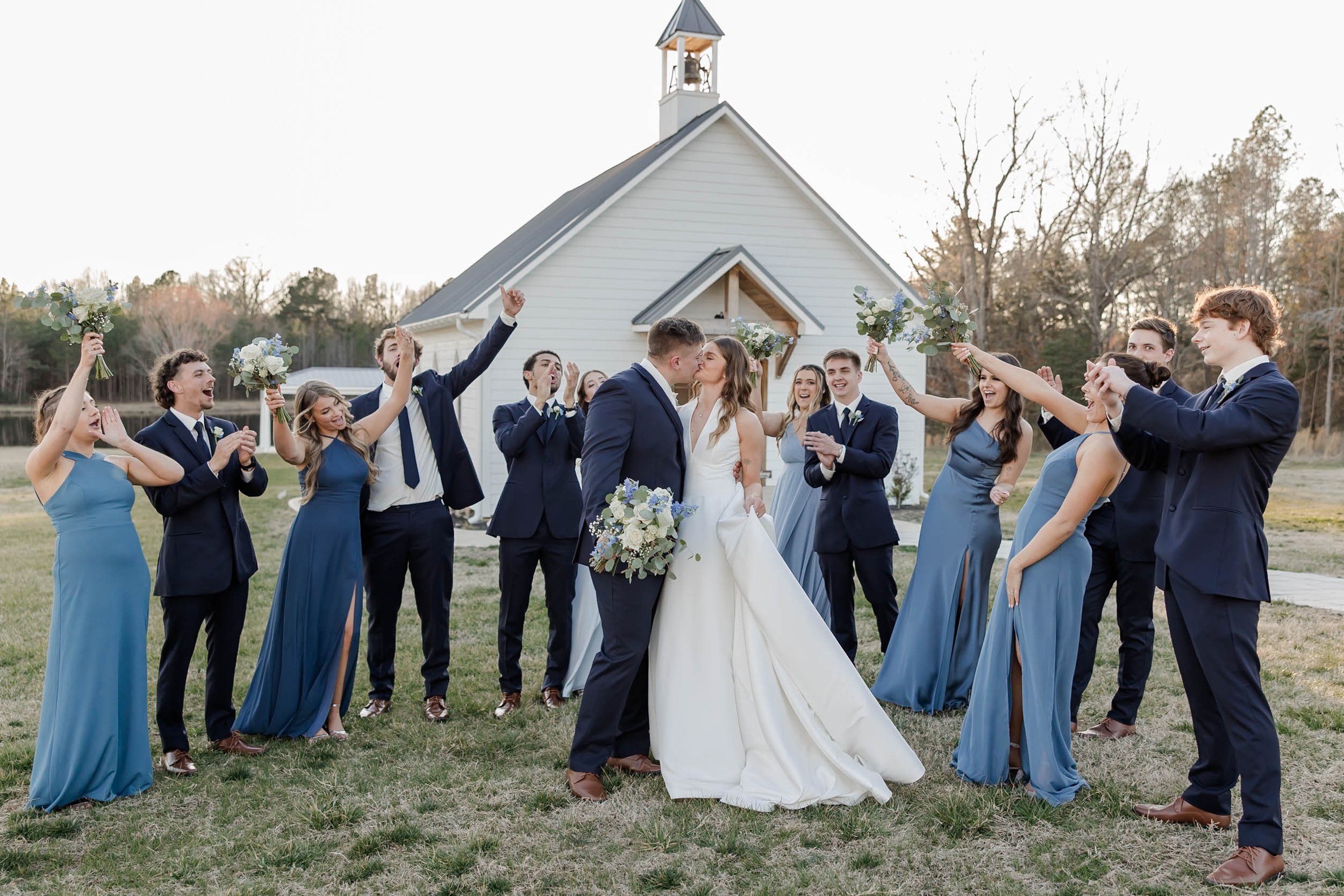 Wedding couple kissing in front of their wedding party outside a small chapel, with bridesmaids and groomsmen cheering and holding bouquets.