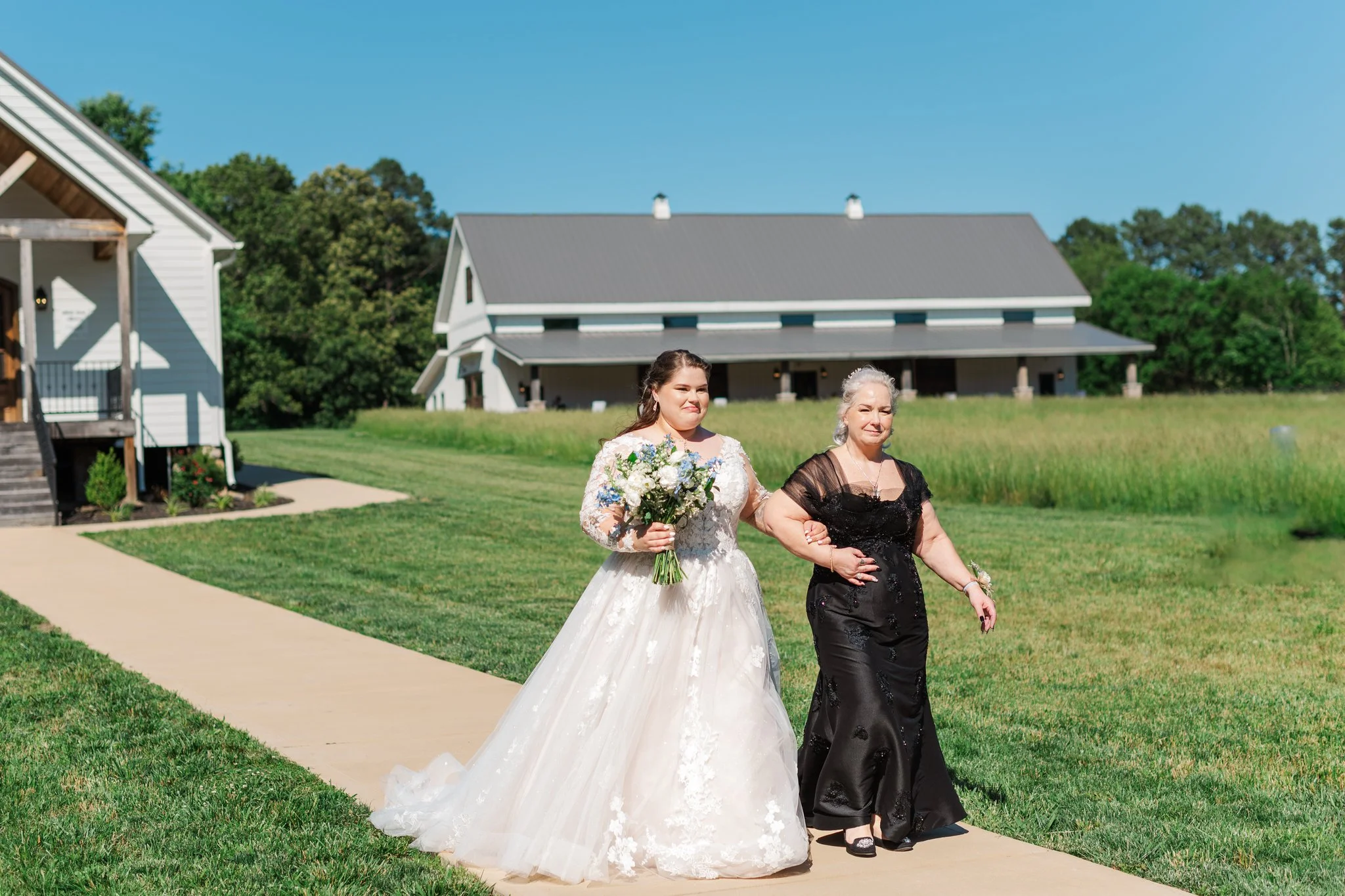 A bride in a white wedding gown holding a bouquet of flowers is walking down the sidewalk with an older woman dressed in a black gown, on a sunny day near a church.