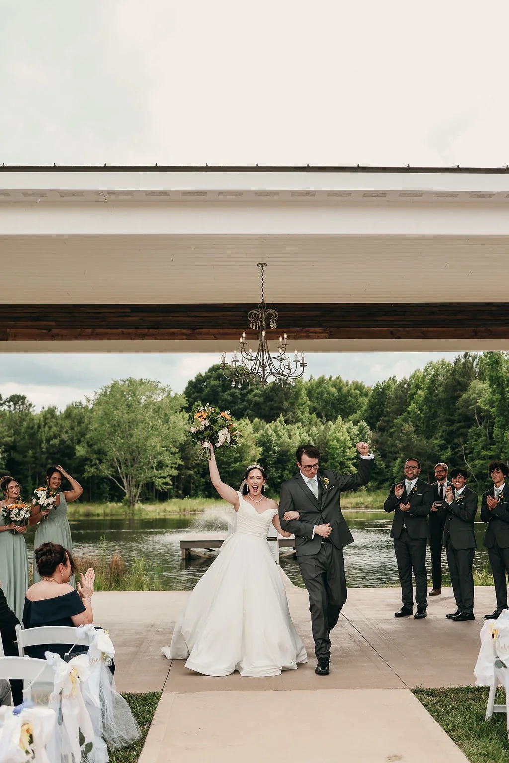 A wedding ceremony by a lake with a bride in a white gown holding a bouquet and a groom in a black suit with a bunch of men and women clapping and celebrating.