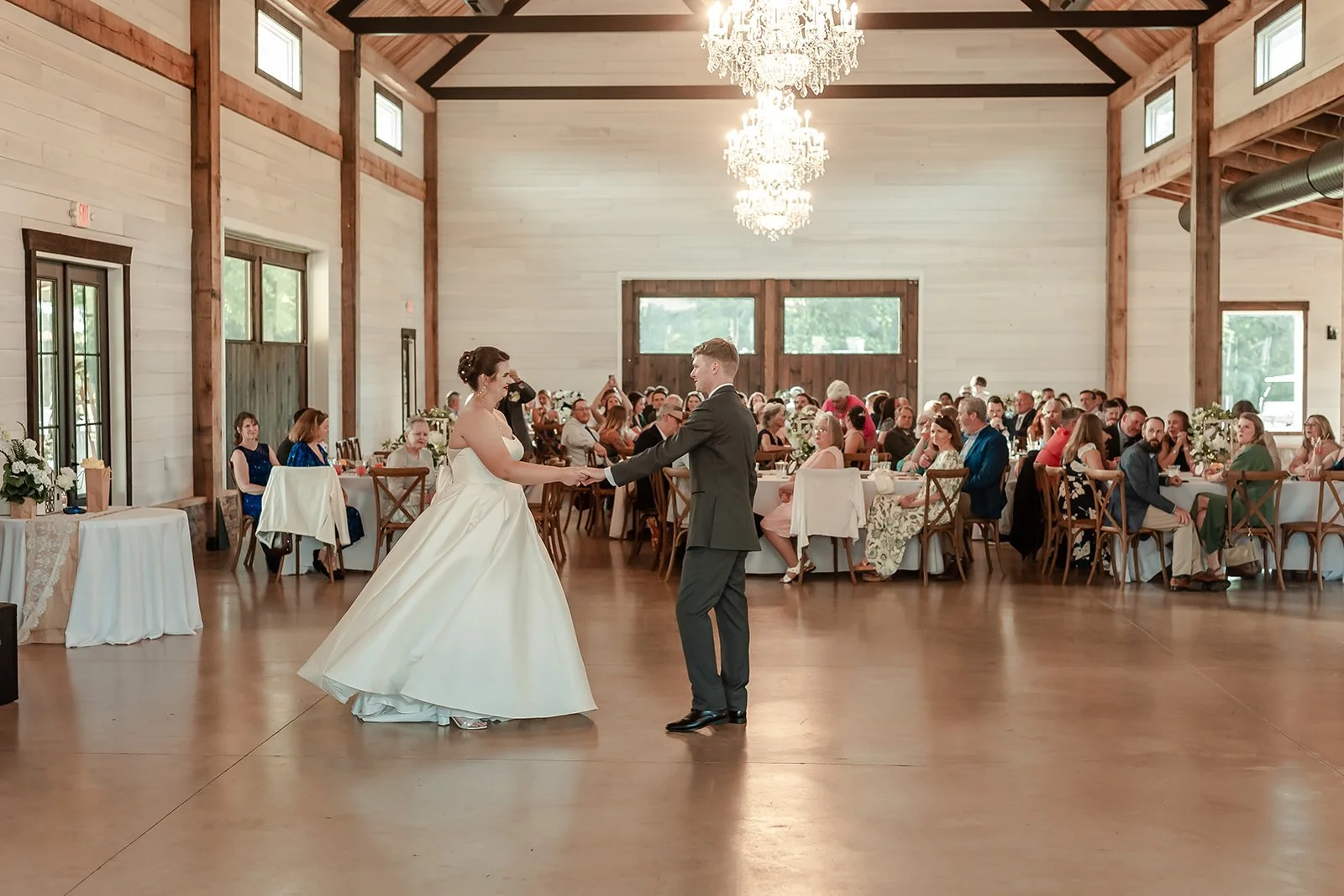 Bride and groom dancing at their wedding reception in a rustic hall with chandeliers and guests seated at tables