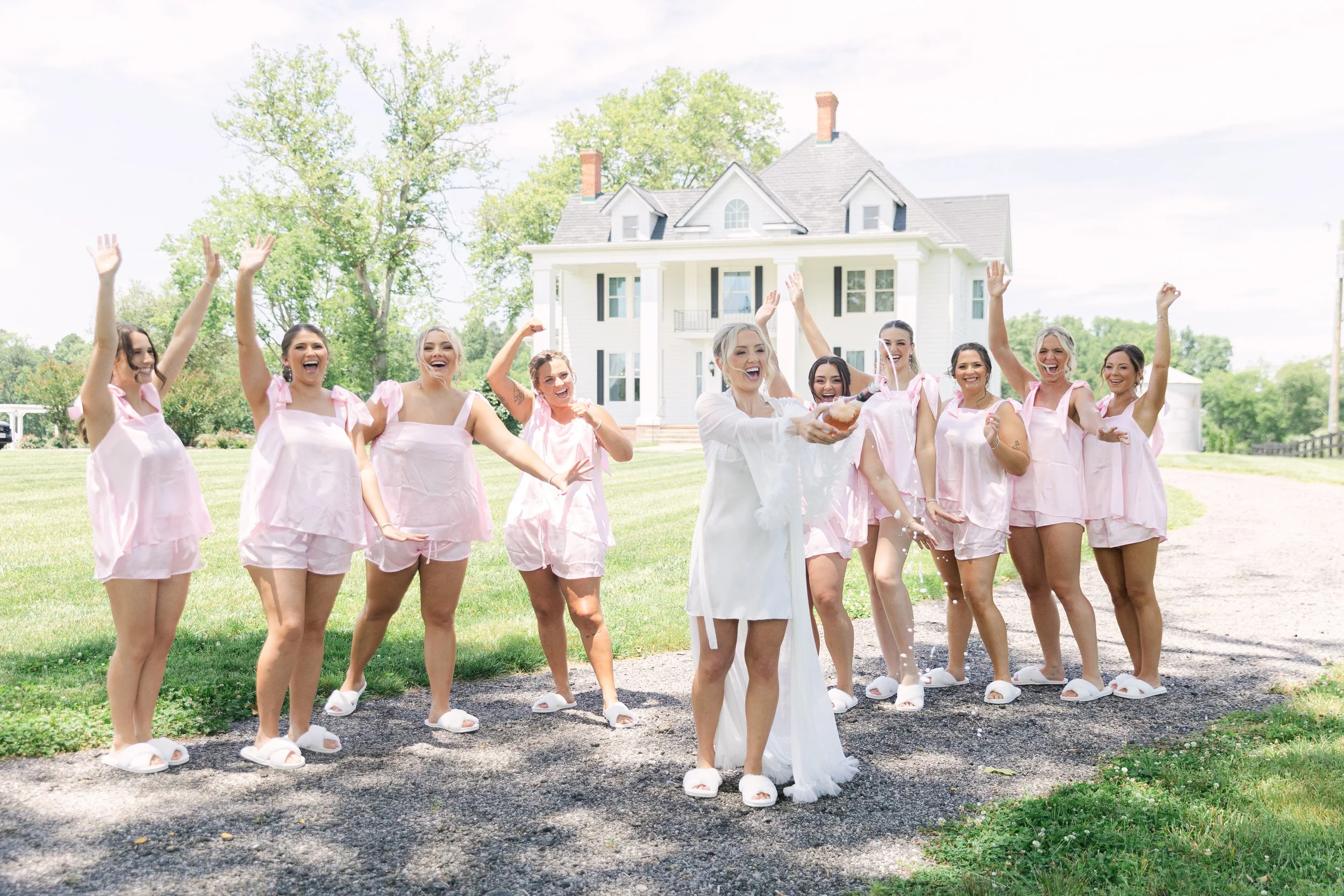 Bride and bridesmaids celebrating outdoors in front of a large white house, with the bride splashing water, all wearing matching pink sleepwear or robes, slippers, on a sunny day.
