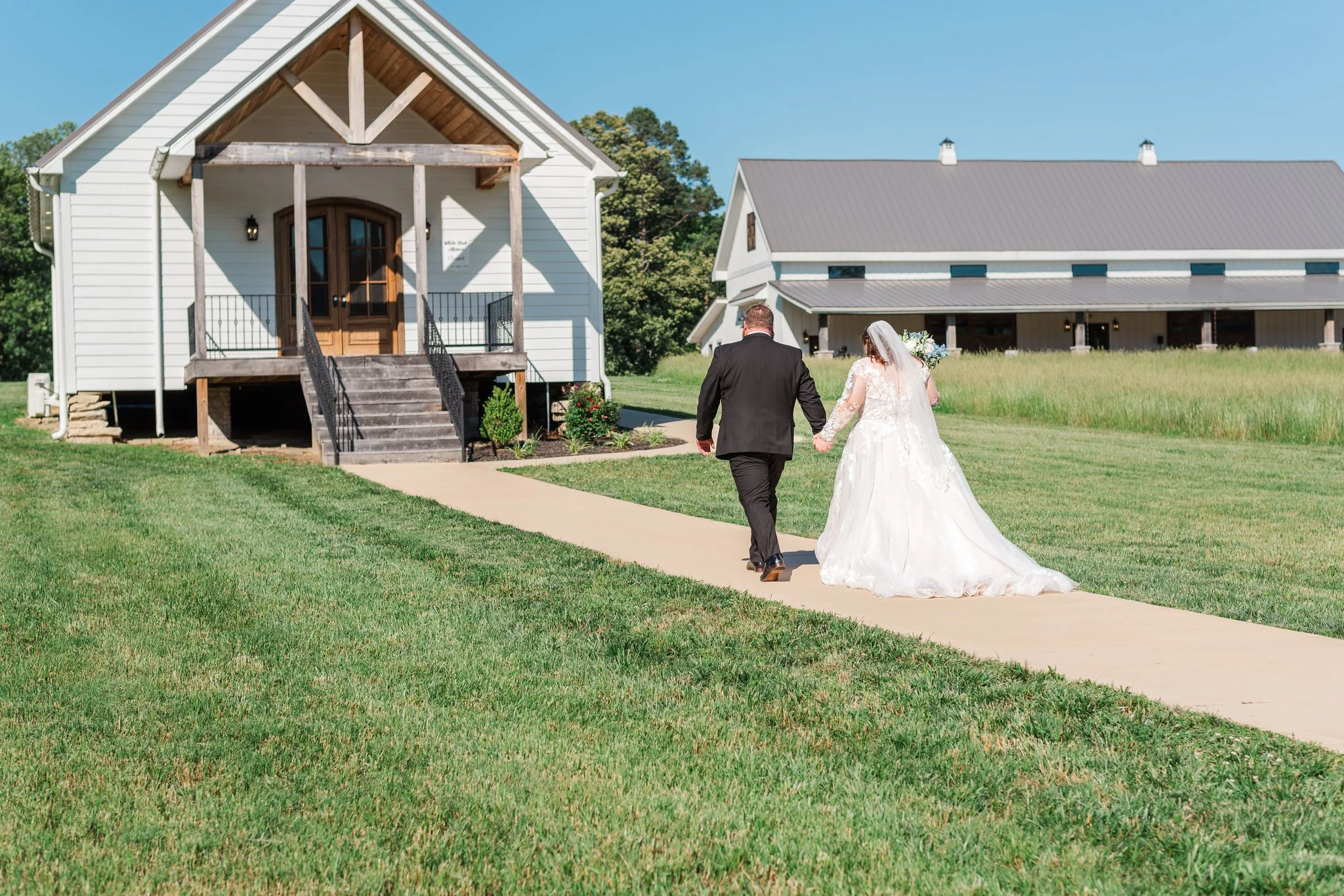 A bride and groom walking hand in hand towards a white house with a porch on a bright sunny day.