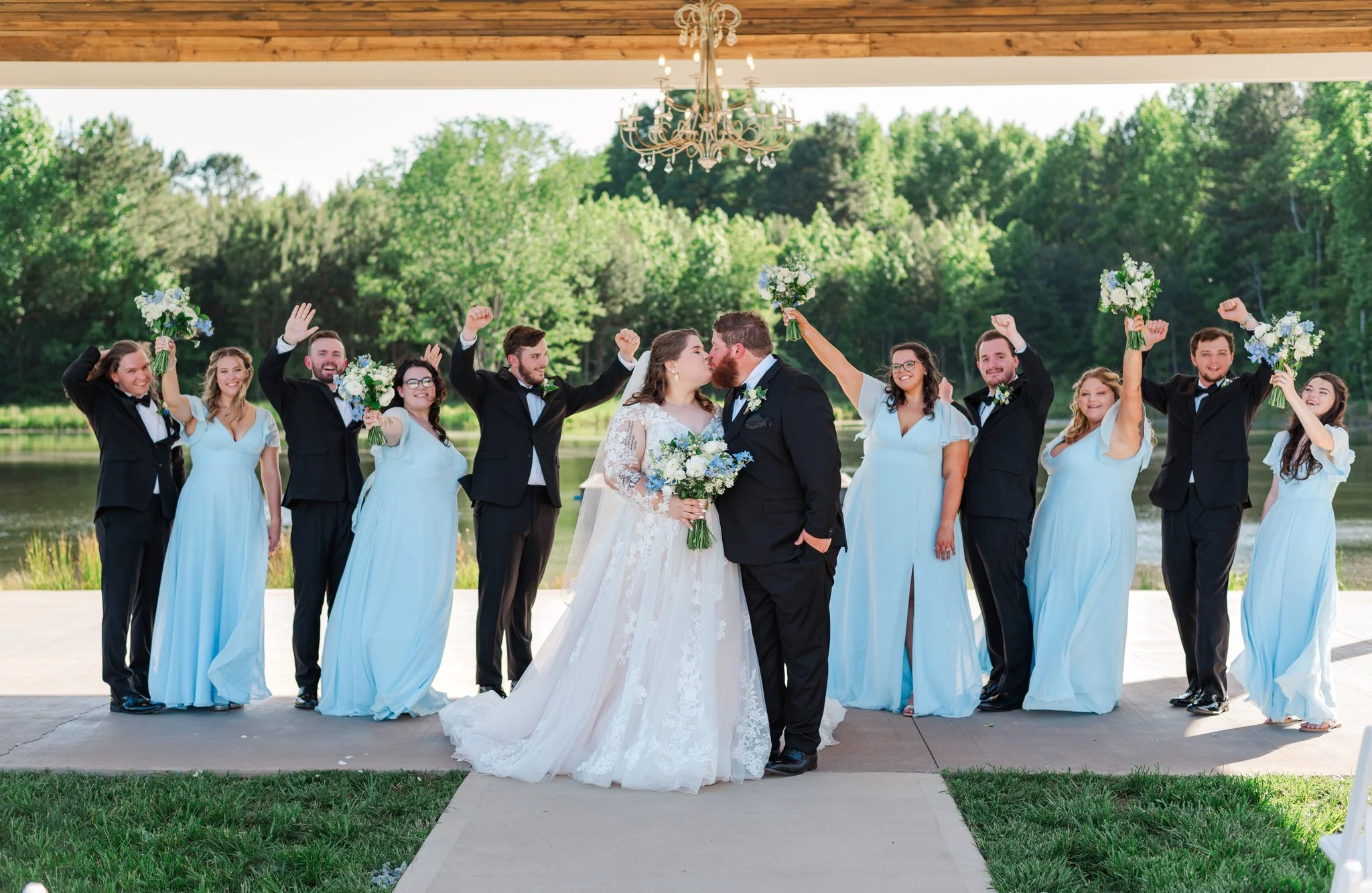Bride and groom kissing under a chandelier with bridesmaids and groomsmen celebrating outdoors near a lake