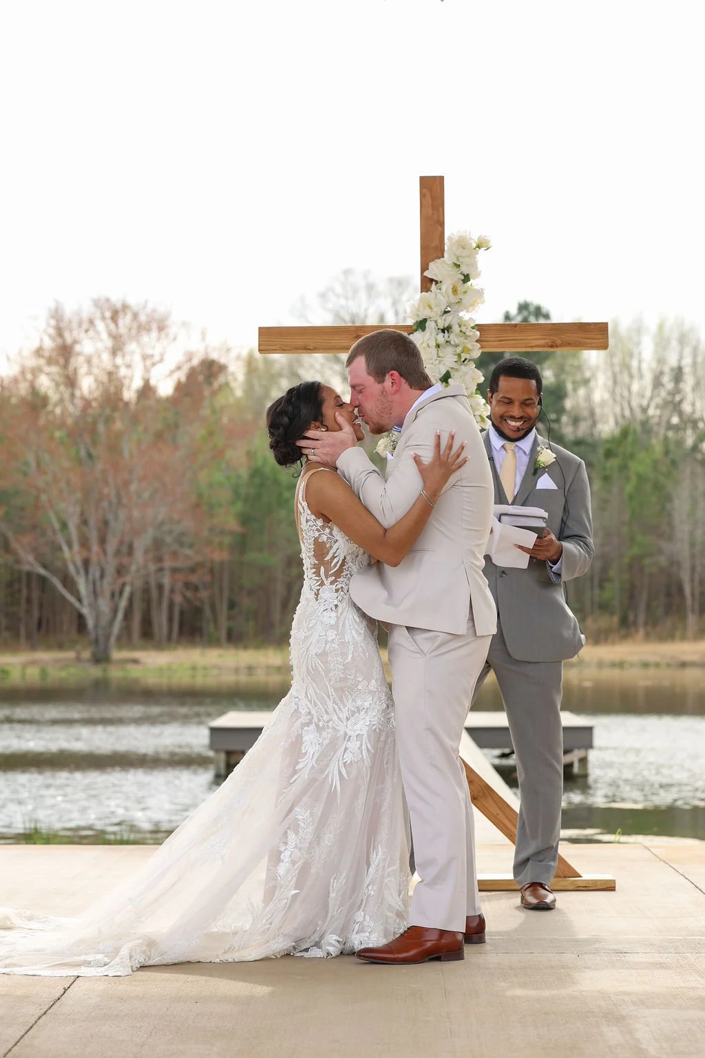 A wedding ceremony outdoors by a lake, with a bride and groom kissing, and an officiant smiling in the background.