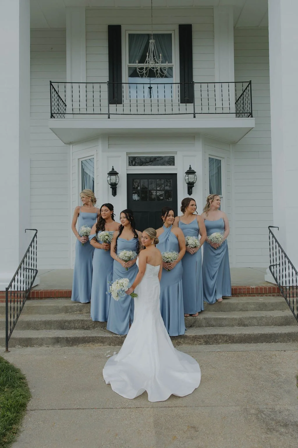 A bride in a white wedding gown standing with her bridal party in front of a white house with black shutters. The bridesmaids are wearing matching blue dresses and holding bouquets of white flowers. They are standing on the steps leading to the house