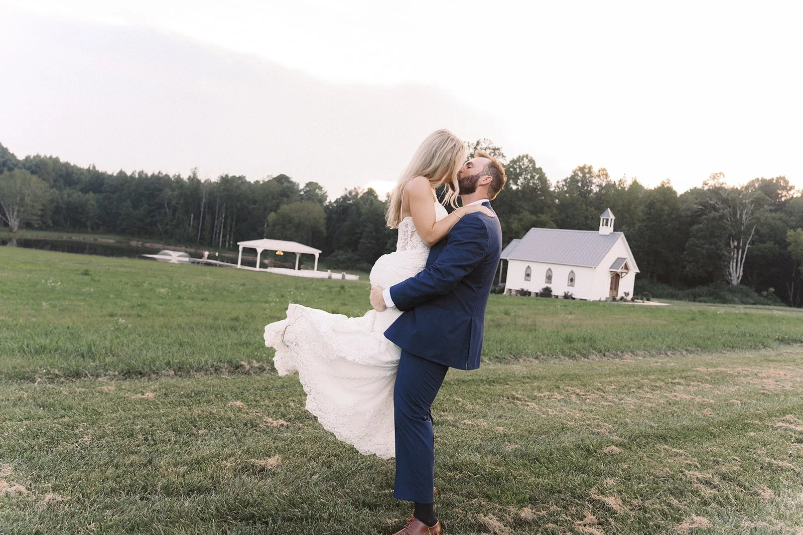 A newlywed couple kissing outdoors on a grassy field. The bride, in a white wedding gown, is being held in the groom's arms. The groom, in a navy blue suit, is lifting her. Background features a small white church, a white gazebo by a pond, and trees