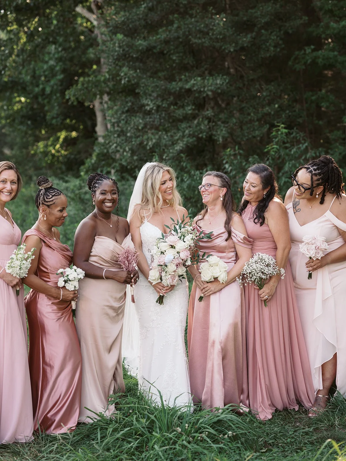 Group of women dressed in pink and white dresses at an outdoor wedding, holding bouquets, standing on grass with trees in the background.