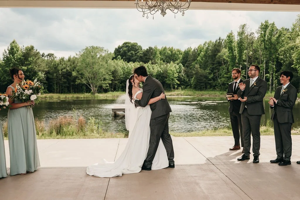 A wedding ceremony outdoors by a lake with a couple kissing, surrounded by bridal party and officiant. The scene is set under a pavilion with a chandelier and lush green trees in the background.