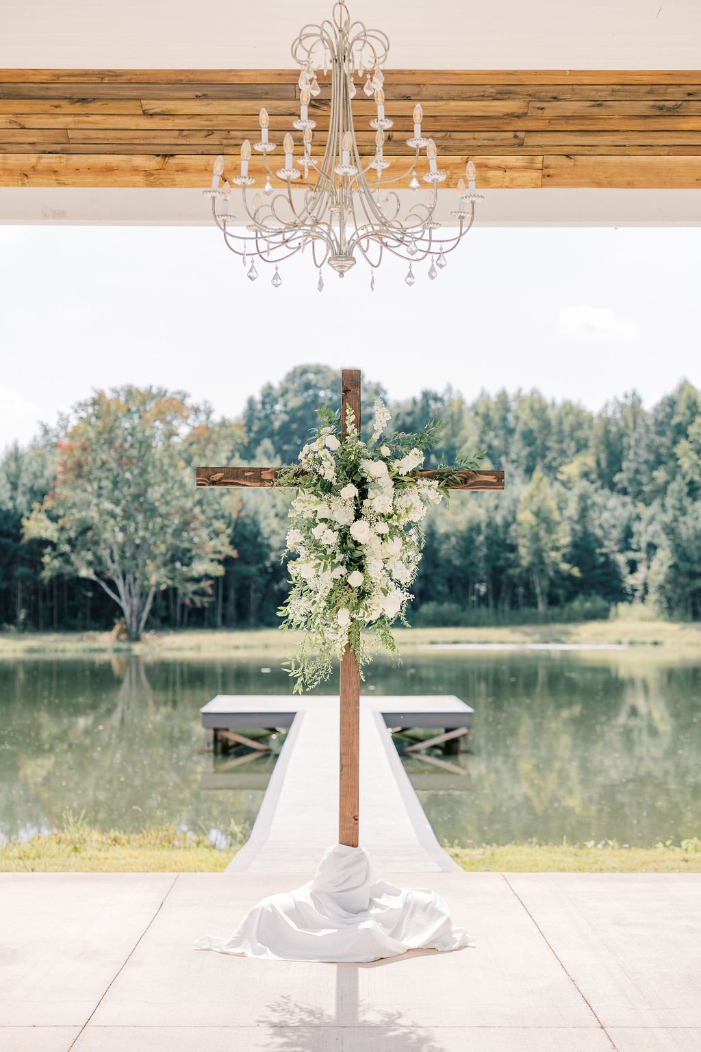 An outdoor wedding ceremony setup with a large wooden cross decorated with white flowers and greenery on a lake dock, a white draped cloth at the base, and a chandelier hanging from a wooden ceiling over the ceremony area.