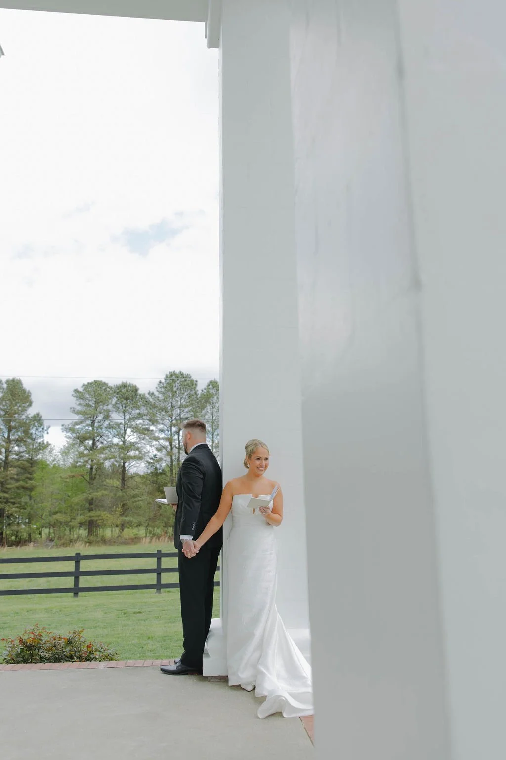 Bride and groom standing outdoors, back to each other, holding hands, with a white building wall between them. The bride is smiling and holding a paper, the groom is holding a cup, and there are trees and a fence in the background.