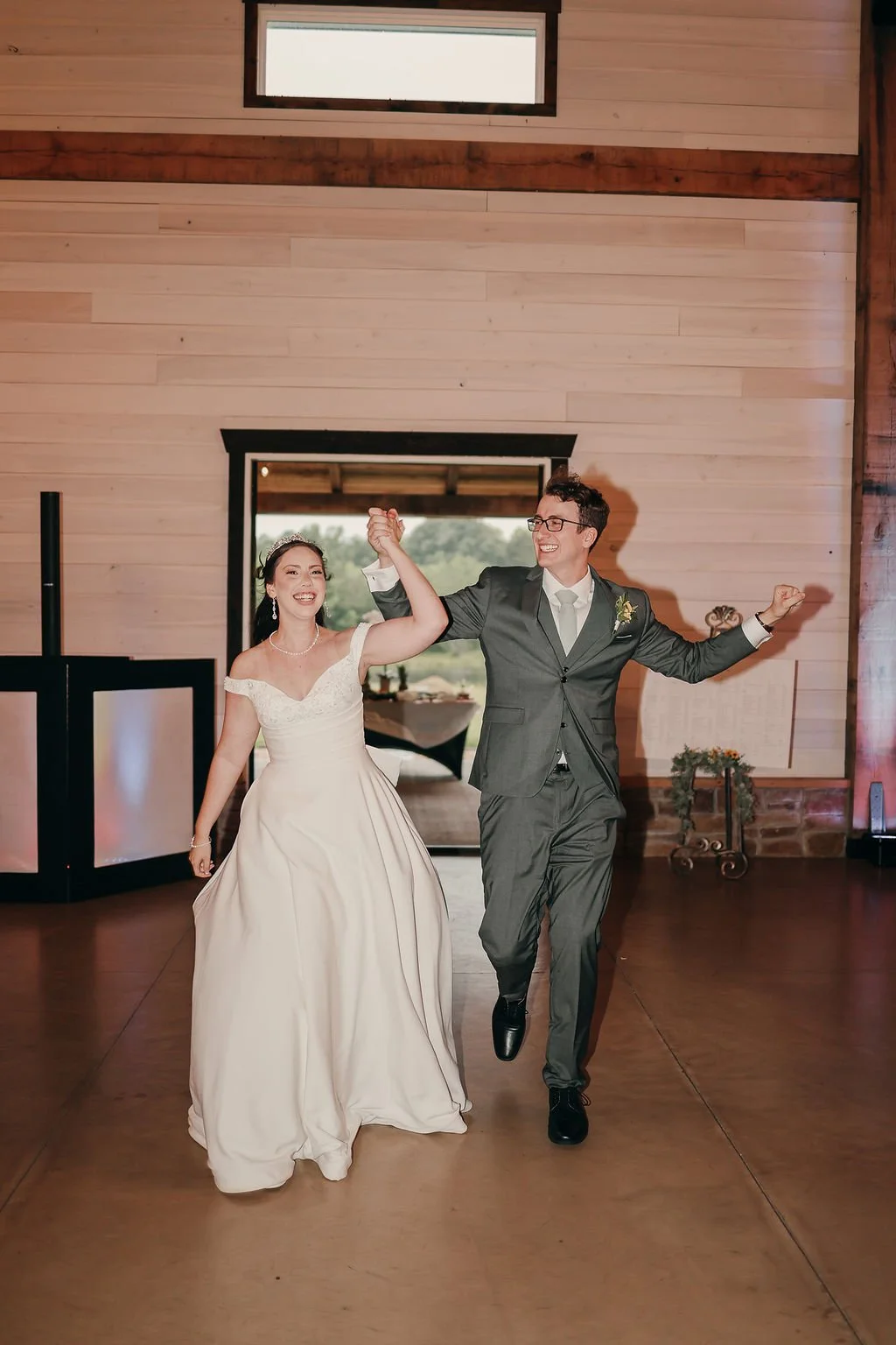 A bride and groom dancing happily at their wedding reception, holding hands and smiling inside a rustic wooden venue.
