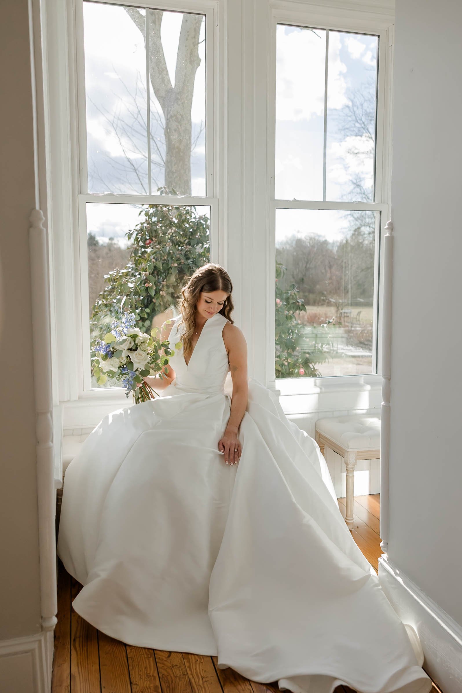 A bride sitting by large windows in a white wedding gown, holding a bouquet of white and purple flowers, with a calm expression.