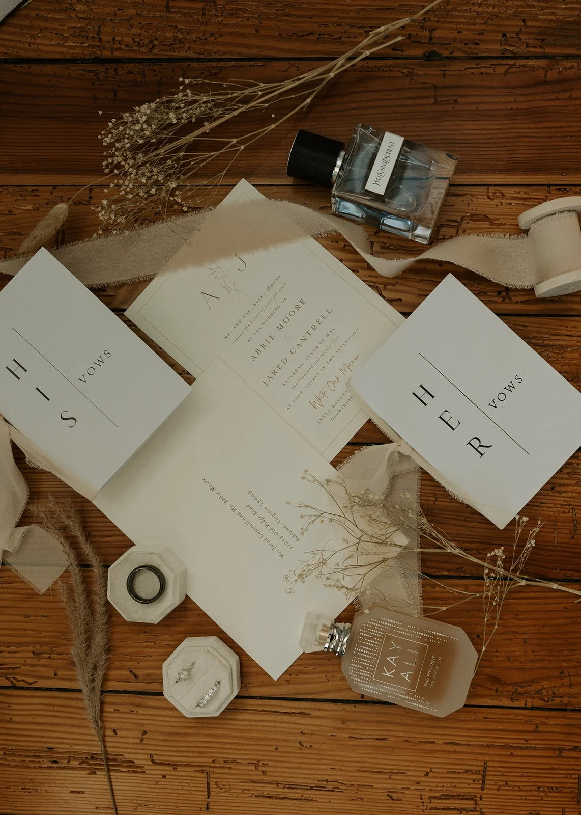 Wedding invitation suite including RSVP card and details card, white rings in a small box, dried flowers, perfume bottles, and fabric ribbons arranged on a wooden surface, with wedding vows printed on paper.