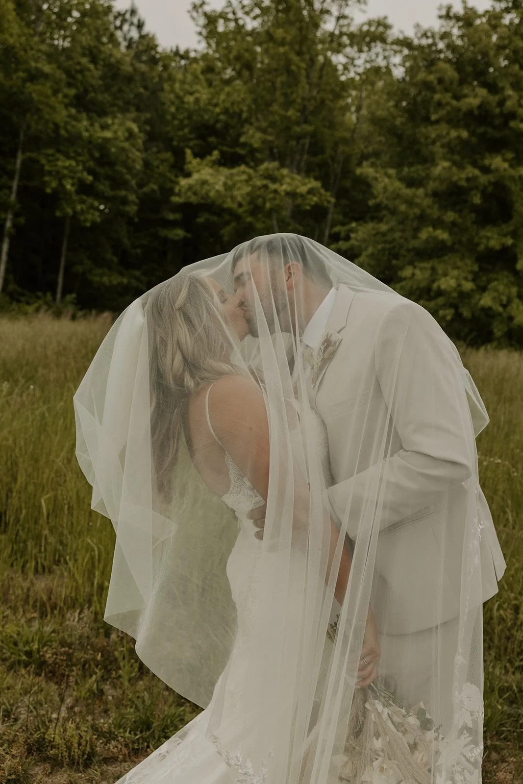 A bride and groom share a kiss under a sheer veil outdoors in a grassy field with trees in the background.
