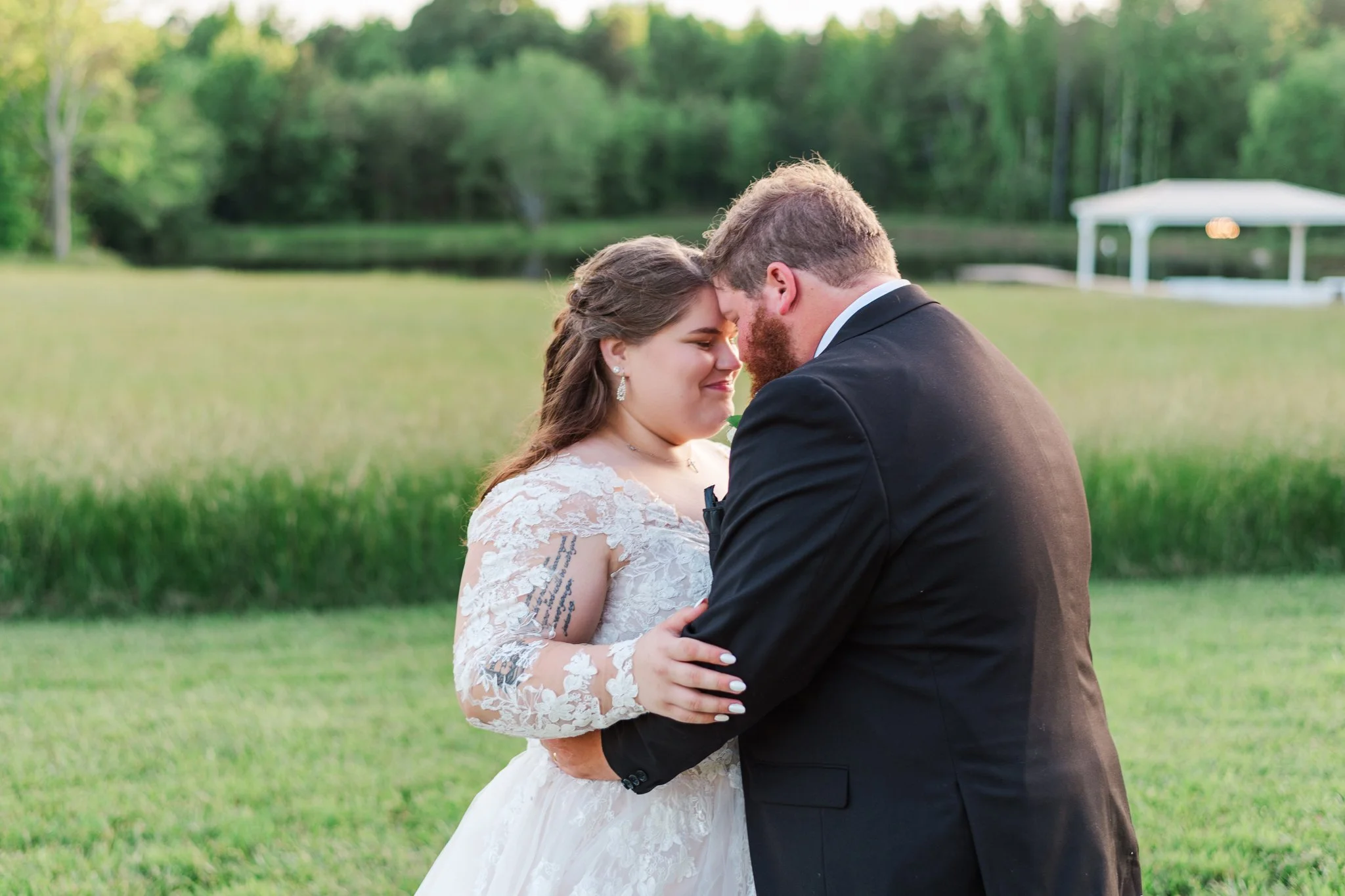 Bride and groom sharing a tender moment outdoors on their wedding day, with a pond and trees in the background.