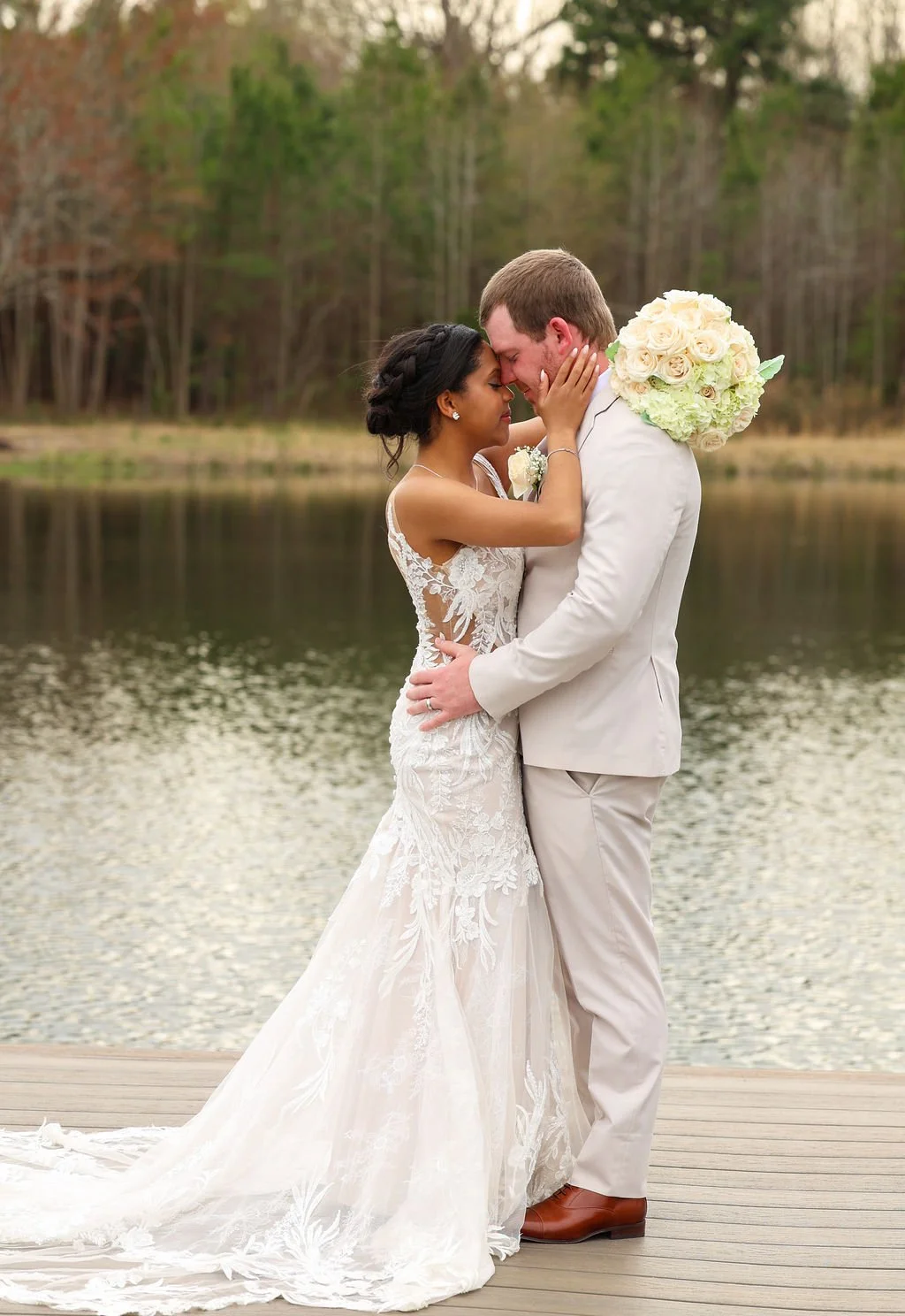 A bride and groom share a romantic moment by a lake with trees in the background, the bride holding a bouquet of white roses on the groom's shoulder.