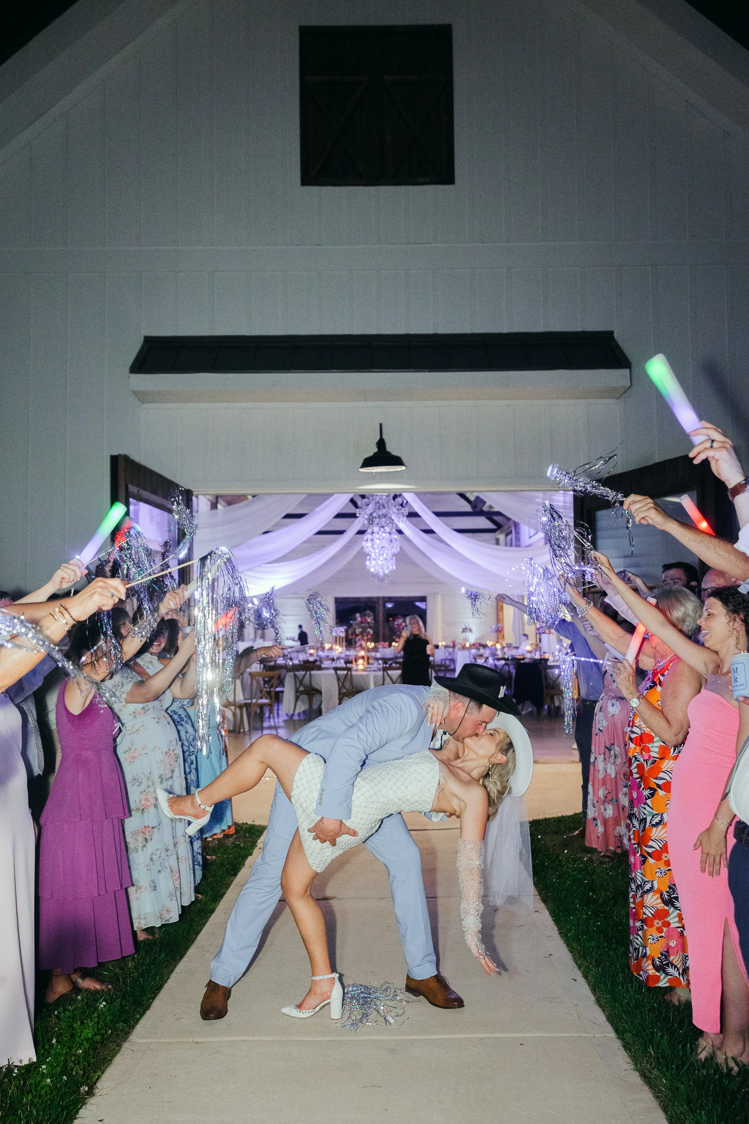 A newlywed couple sharing a kiss during their wedding celebration, with guests on either side holding glow sticks and silver decorations, in a decorated outdoor reception area at night.