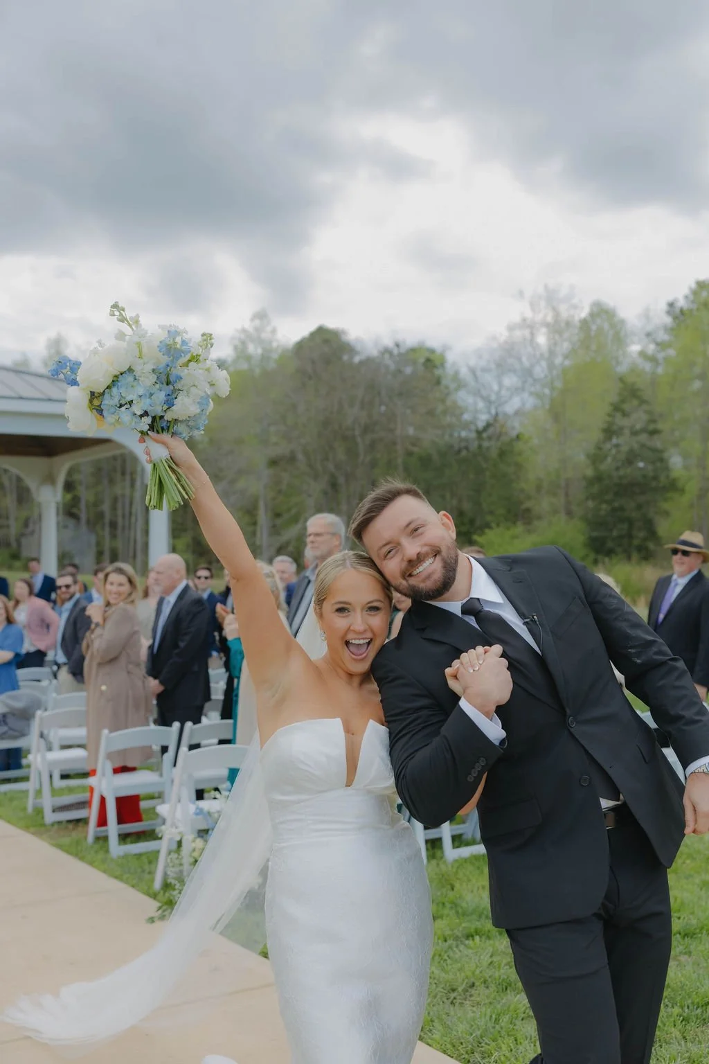 Happy bride and groom celebrating at outdoor wedding, woman holding bouquet, guests in background.