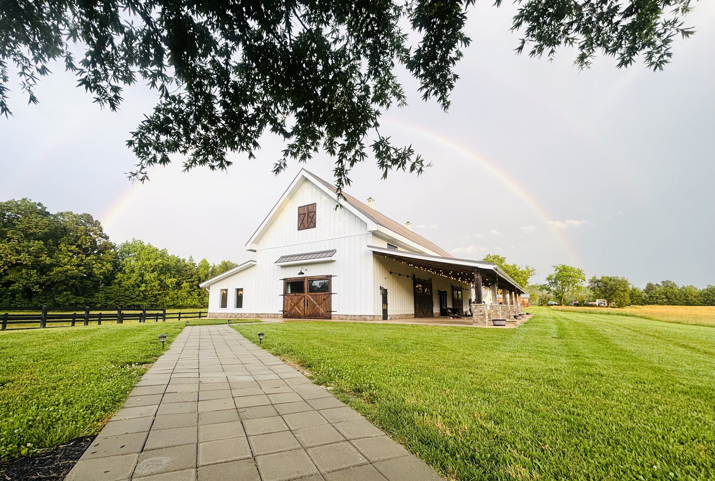 A white barn with a brown roof and sliding doors on a large grassy field, with a paved walkway leading to it. There is a rainbow in the sky above the barn amidst clouds, and tree branches framing the top of the image.