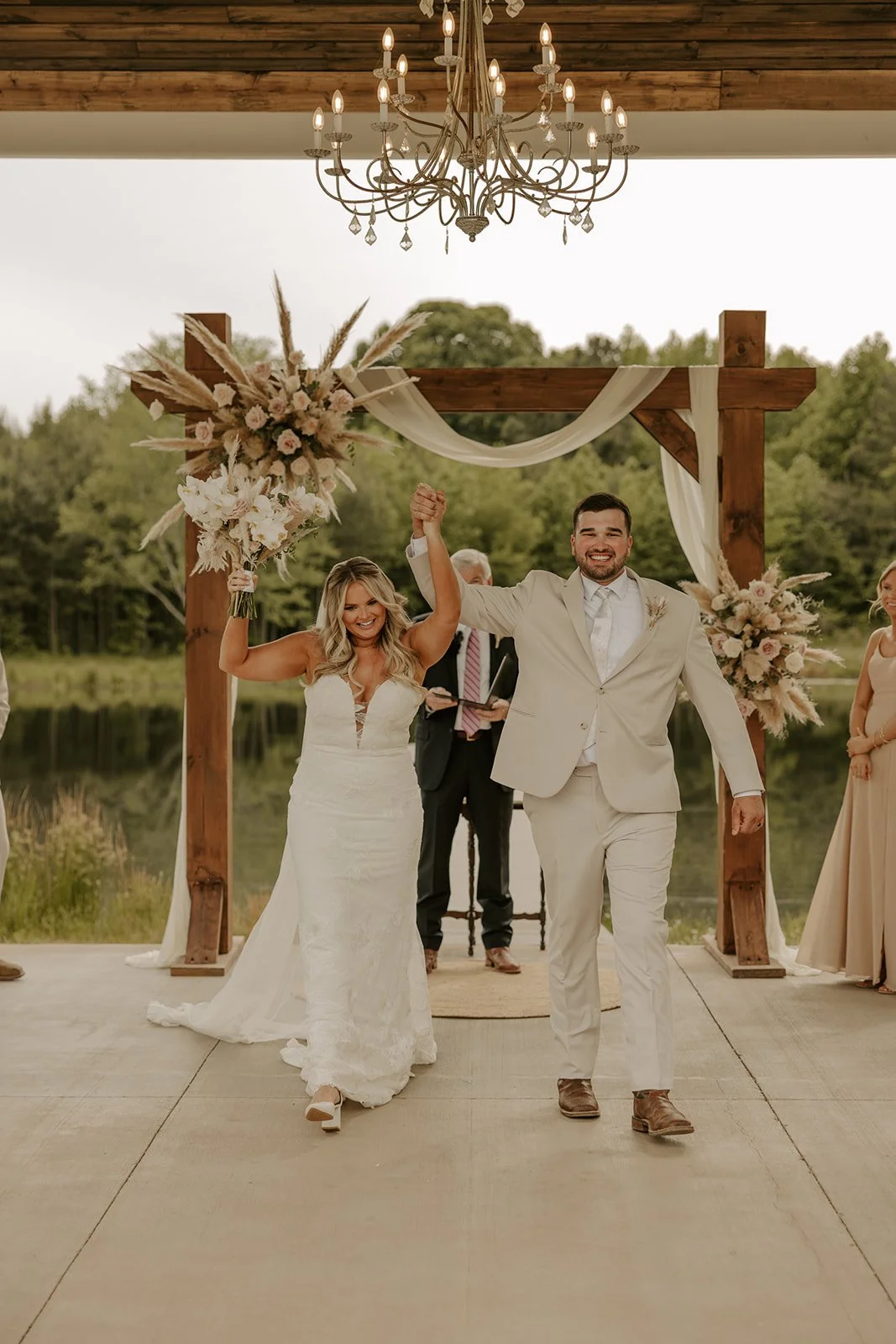 A newlywed couple holding hands and smiling as they walk down the aisle outdoors under a wooden arch with floral decorations during their wedding ceremony.
