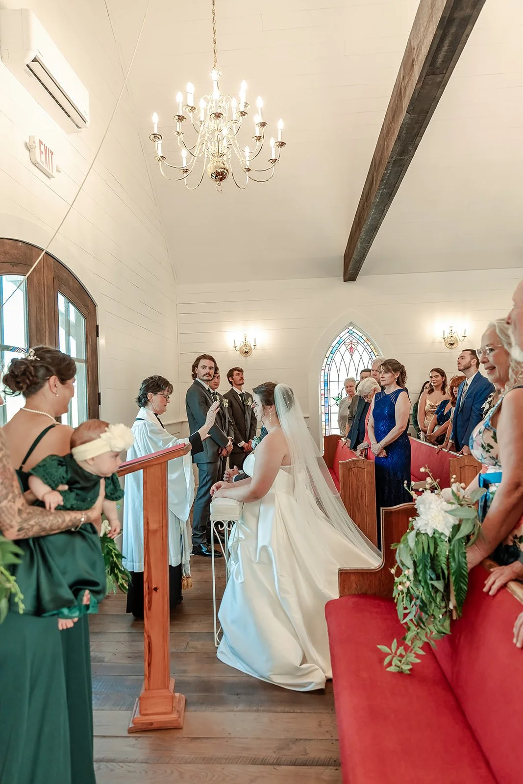 A wedding ceremony in a small church with the bride, groom, and celebrated officials. The church has white walls, a chandelier, and stained glass windows.
