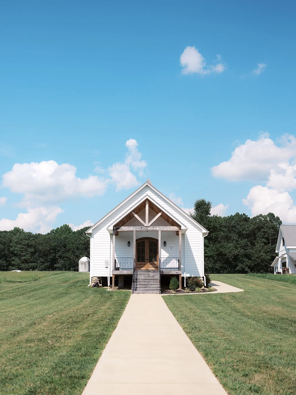 White house with a porch, wooden stairs, and a front door, set on green grass with a sidewalk leading up to it, under a blue sky with clouds.