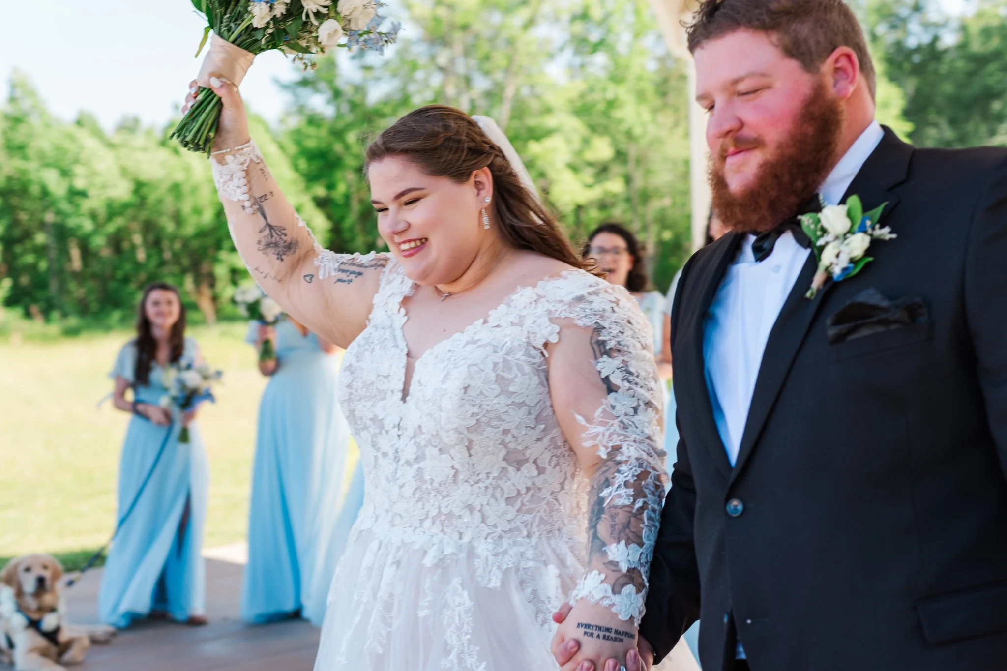 A bride and groom holding hands at their outdoor wedding ceremony, with the bride holding a bouquet, smiling, and the groom dressed in a tuxedo with a boutonniere, surrounded by bridesmaids and greenery.