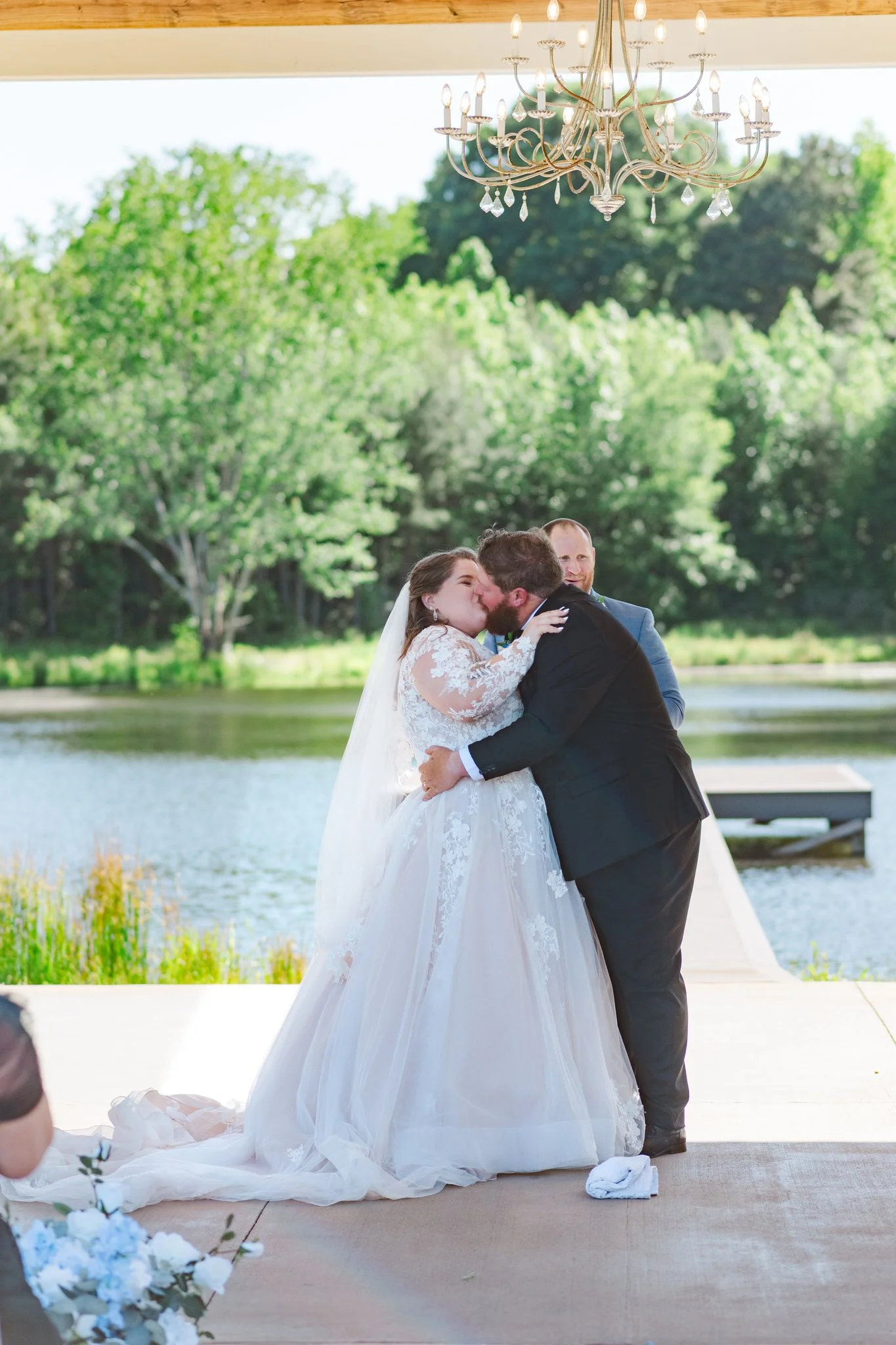 Bride and groom kiss during wedding ceremony outdoors near a lake with greenery, under a chandelier.