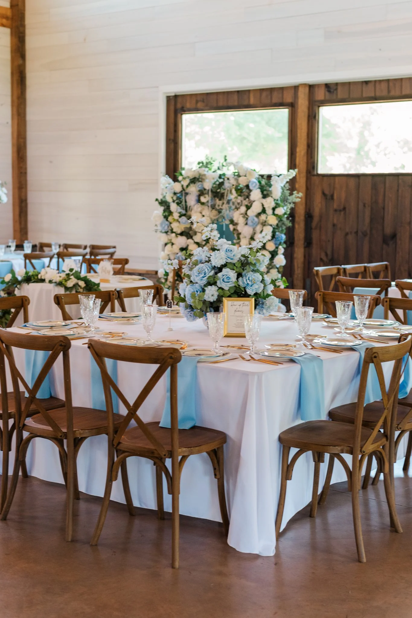 A decorated indoor dining table at a special event, featuring white tablecloths, floral centerpieces in shades of white and light blue, and set with glasses and plates, with wooden chairs surrounding the table and a rustic wooden wall with windows in