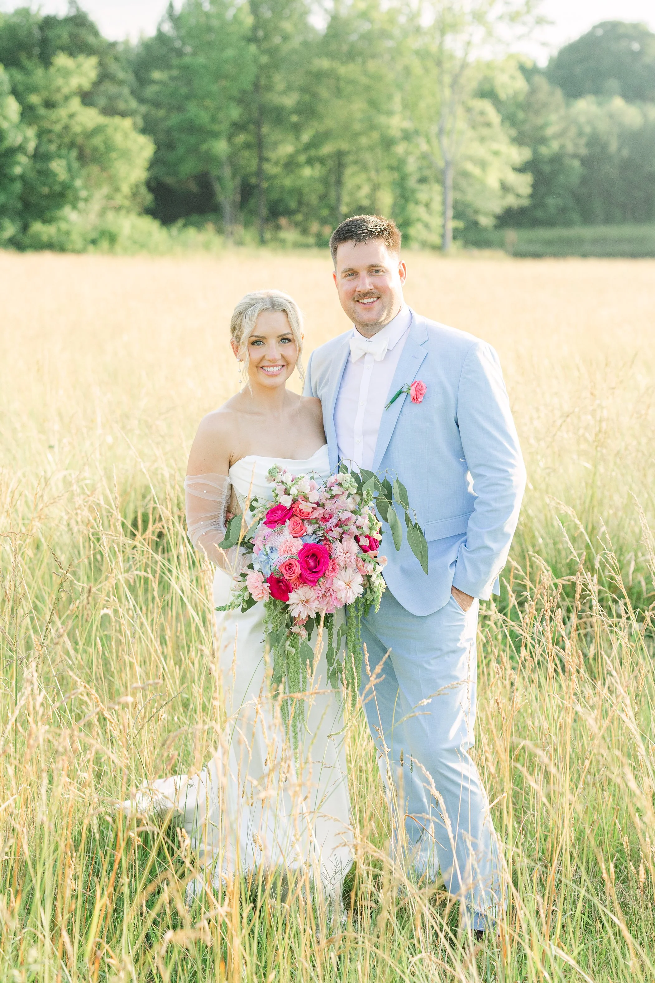 A smiling bride and groom standing in a field of tall grass with a forested background. The bride is holding a large bouquet of pink and white flowers, and the groom is wearing a light blue suit with a pink flower boutonniere.