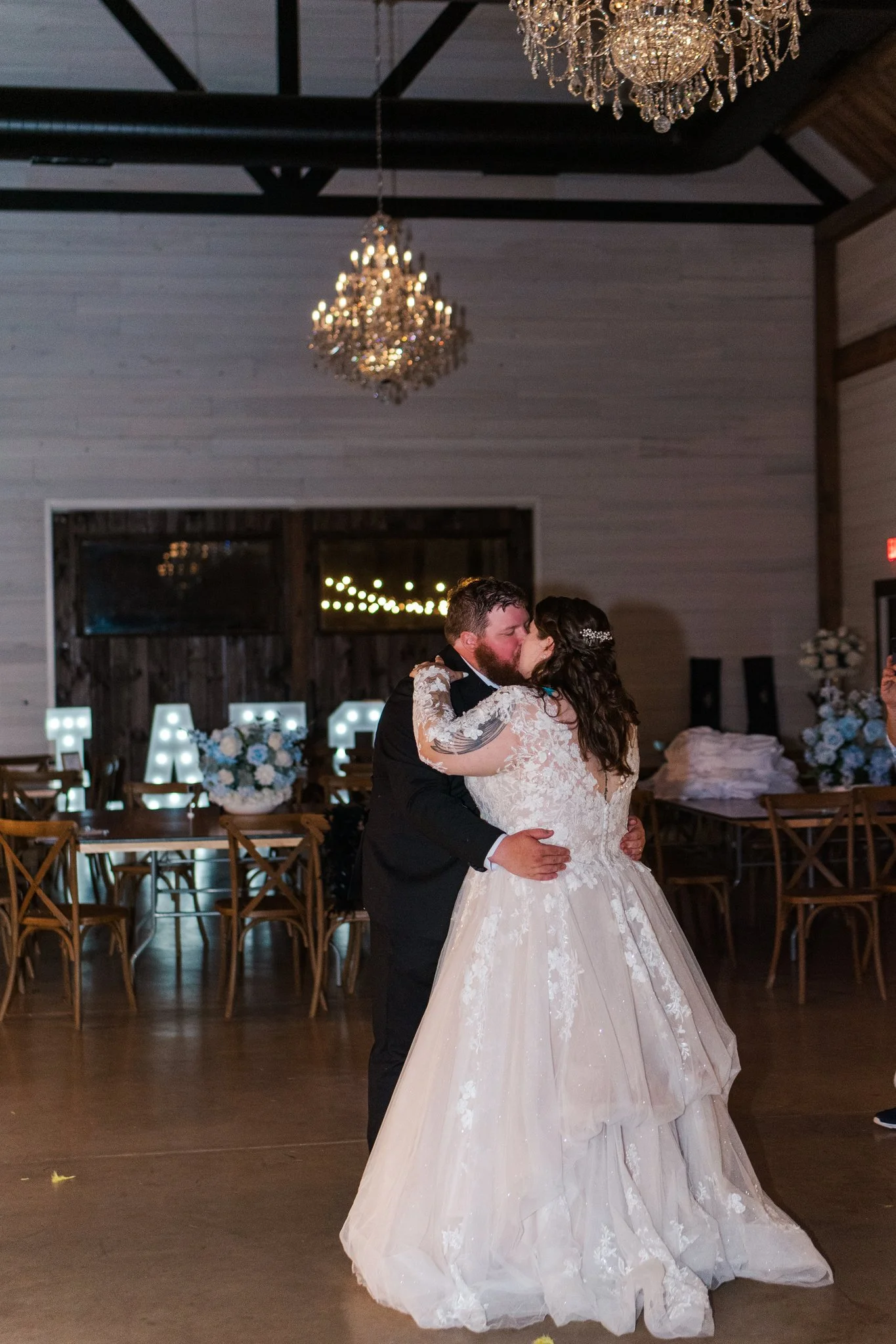 A bride and groom share a kiss while dancing at their wedding in a decorated reception hall with chandeliers and illuminated letters.