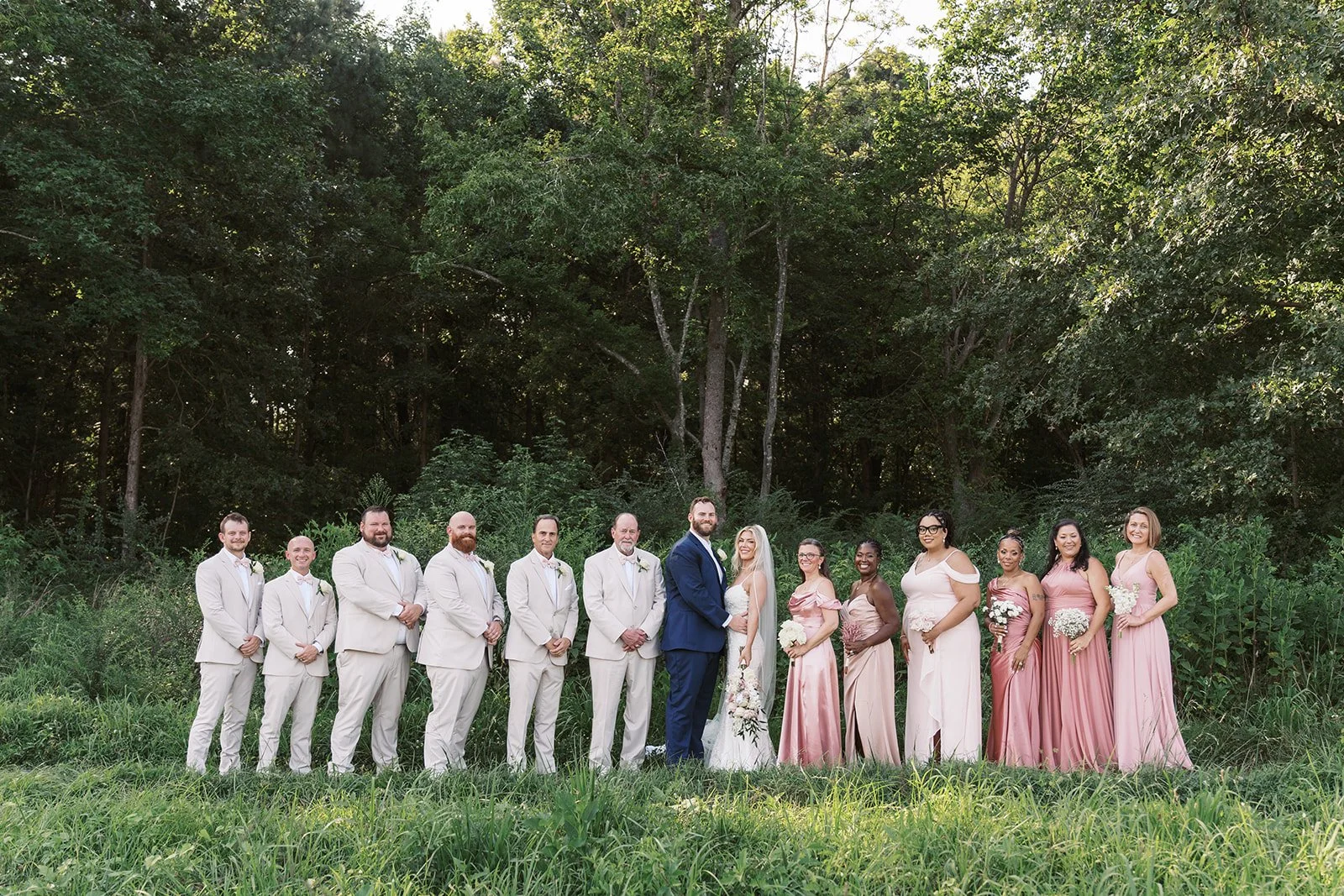 A group of wedding guests standing outdoors on grass with trees in the background, including men in beige suits and women in pink dresses, with the bride and groom in the center.