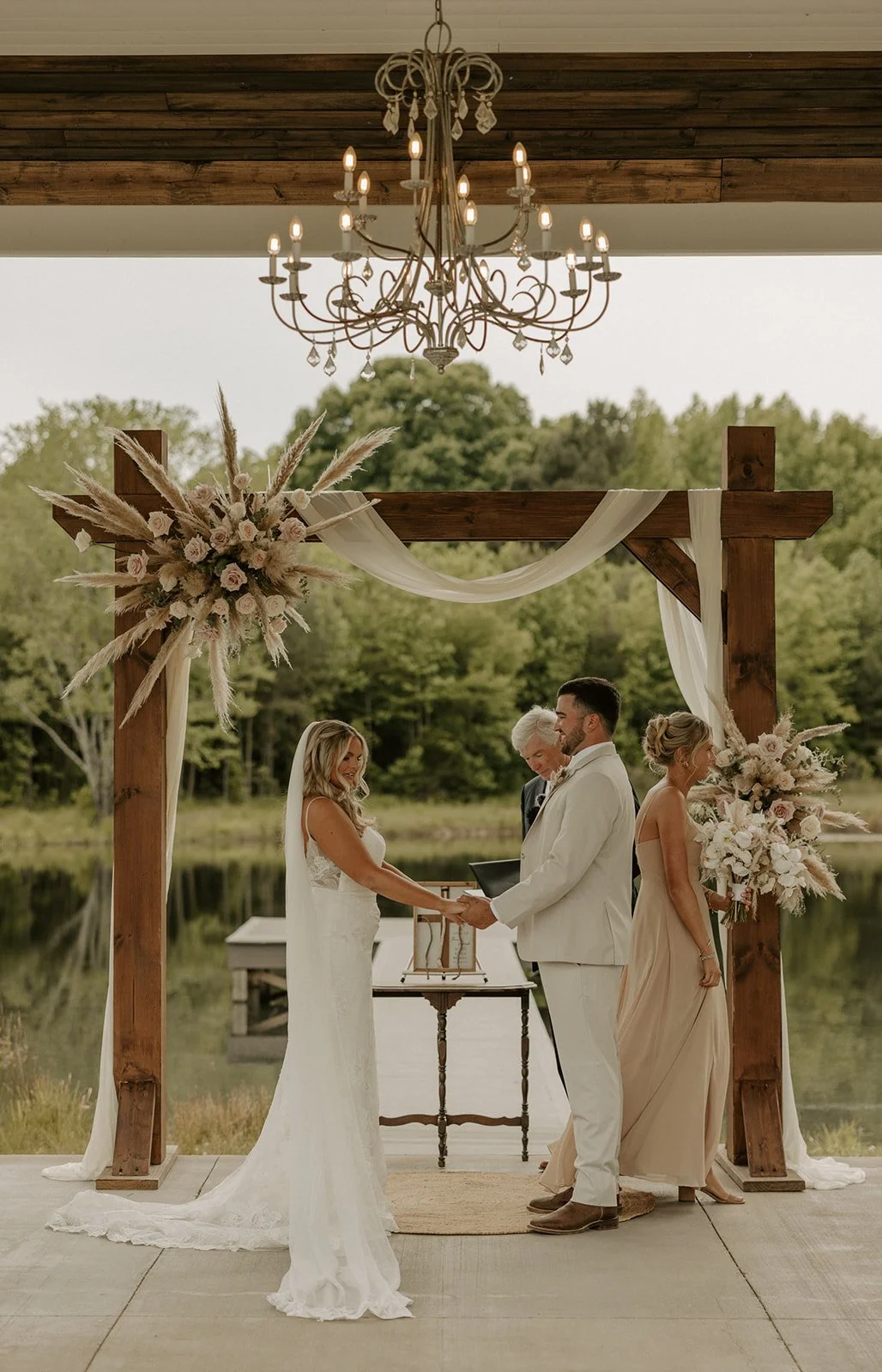 A couple getting married outdoors, facing each other and holding hands, with an officiant standing behind a small table. They are under a wooden arch decorated with flowers and drapery, near a lake, with trees in the background. An elegant chandelier