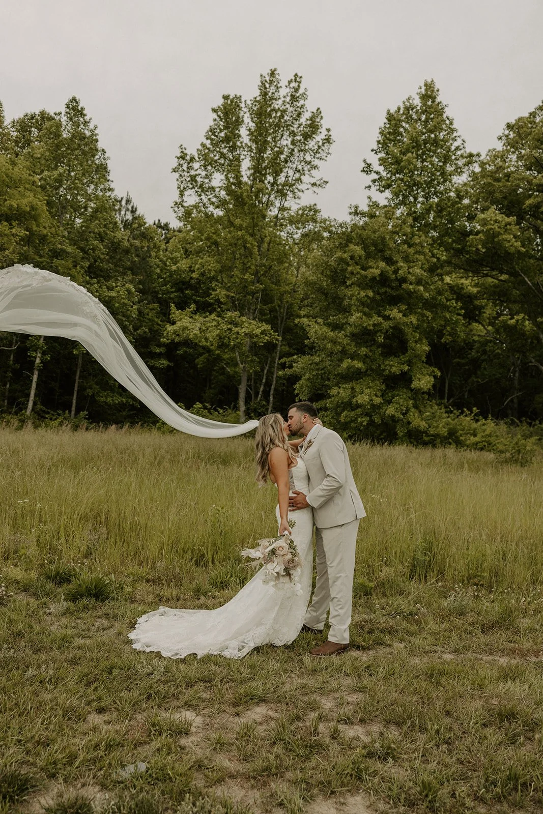 A bride and groom sharing a kiss outdoors in a grassy field with trees in the background, the bride holding a bouquet of flowers, with a flowing veil trailing in the air.