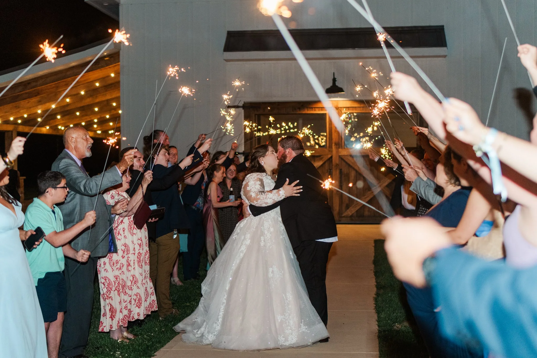 Bride and groom kissing surrounded by guests holding sparklers at night outside a wedding venue.