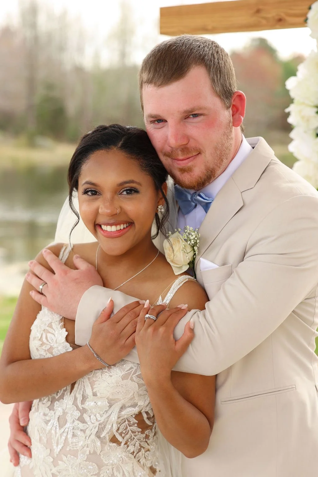 A newlywed couple embraces outdoors near a lake, with the bride in a white lace wedding dress and the groom in a beige suit and blue bow tie, both smiling happily.