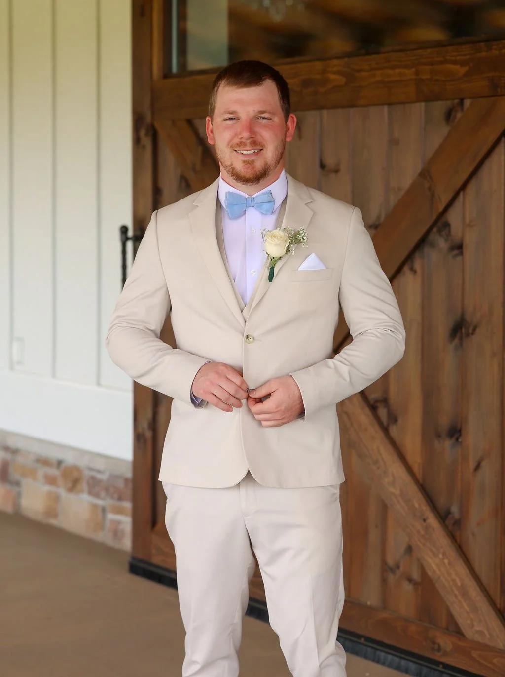 A man in a light beige suit with a white shirt and light blue bow tie, standing in front of a wooden barn door, smiling and adjusting his suit.