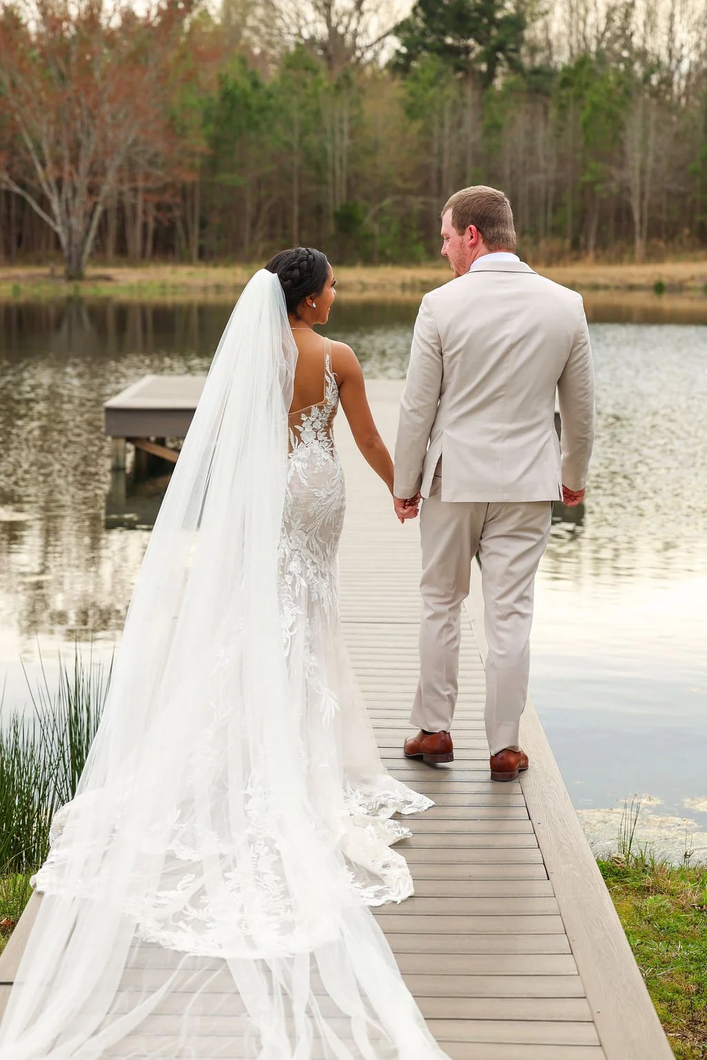 A bride and groom holding hands on a wooden dock by a lake with trees in the background.