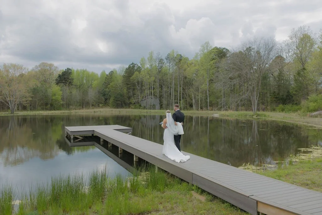 A bride and groom standing on a wooden dock by a pond, surrounded by trees and a cloudy sky.
