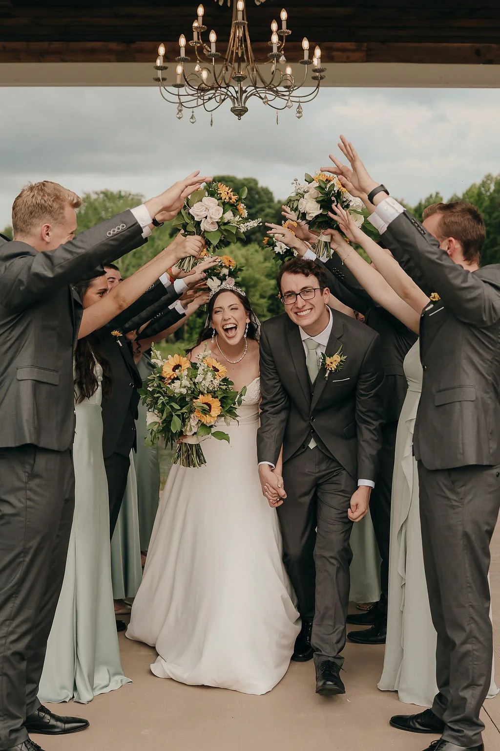 A bride and groom walking hand in hand, smiling, surrounded by friends and family holding bouquets above their heads in an outdoor wedding celebration.