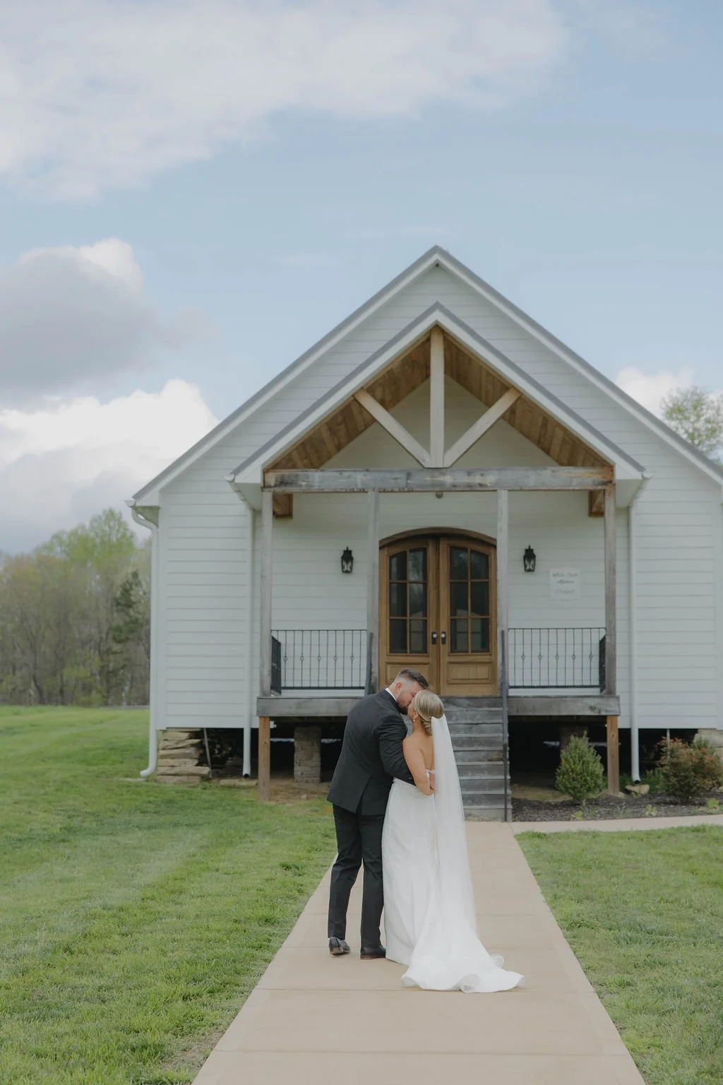 A newlywed couple in wedding attire sharing a moment outside a white house with a wooden door and porch, green lawn, and cloudy sky.