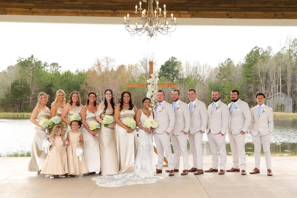 A wedding party posed outdoors near a lake, with a cross and floral arrangement in the background, under a large chandelier hanging from a wooden ceiling.